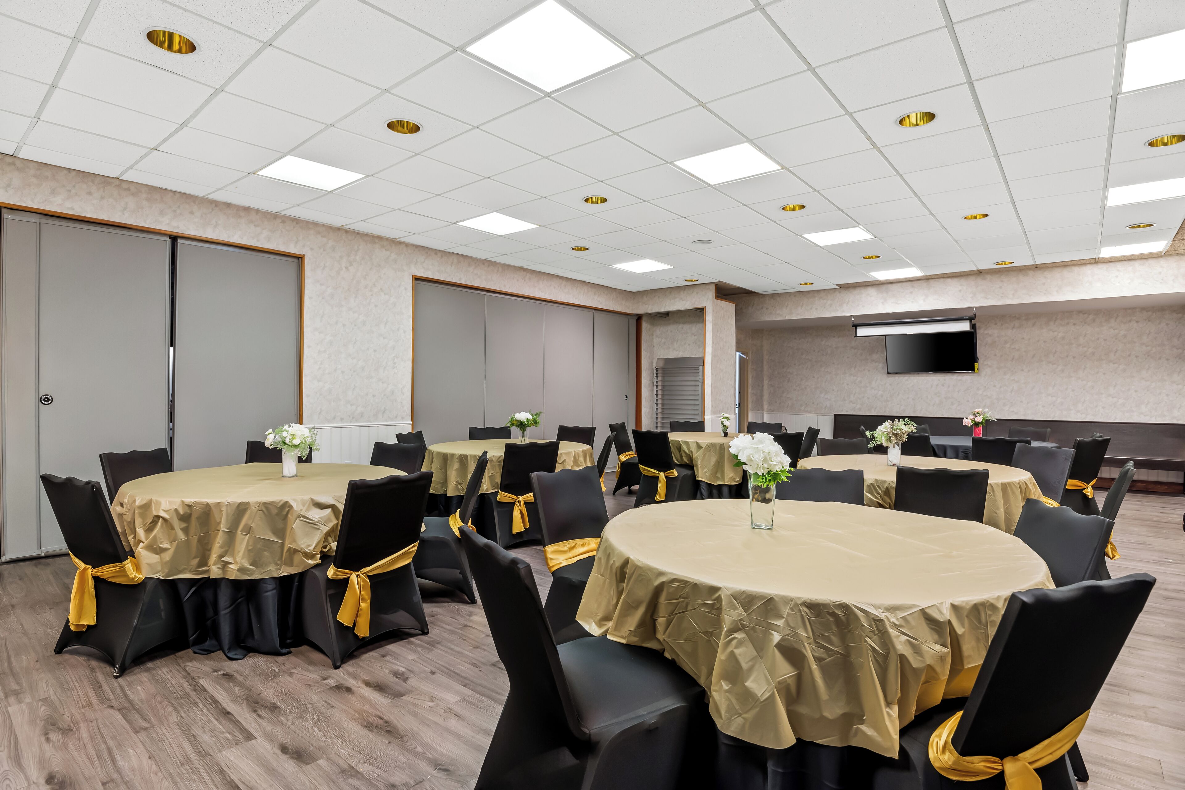 A conference room with multiple round tables, black chairs with yellow sashes, and a flat-screen TV mounted on the wall.