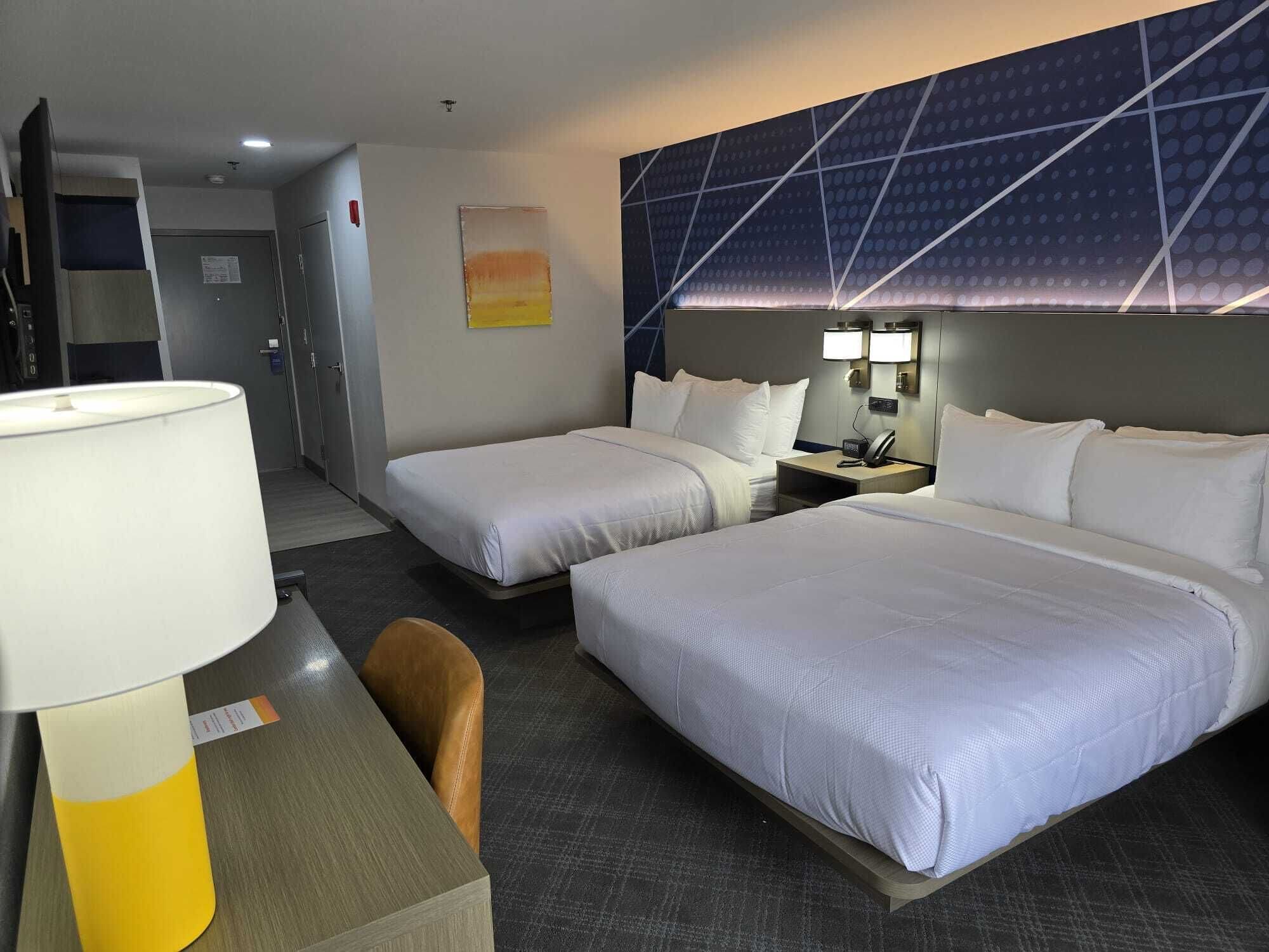 Habitación de hotel con dos camas, un escritorio con lámpara, una silla y una pared con diseño geométrico.
