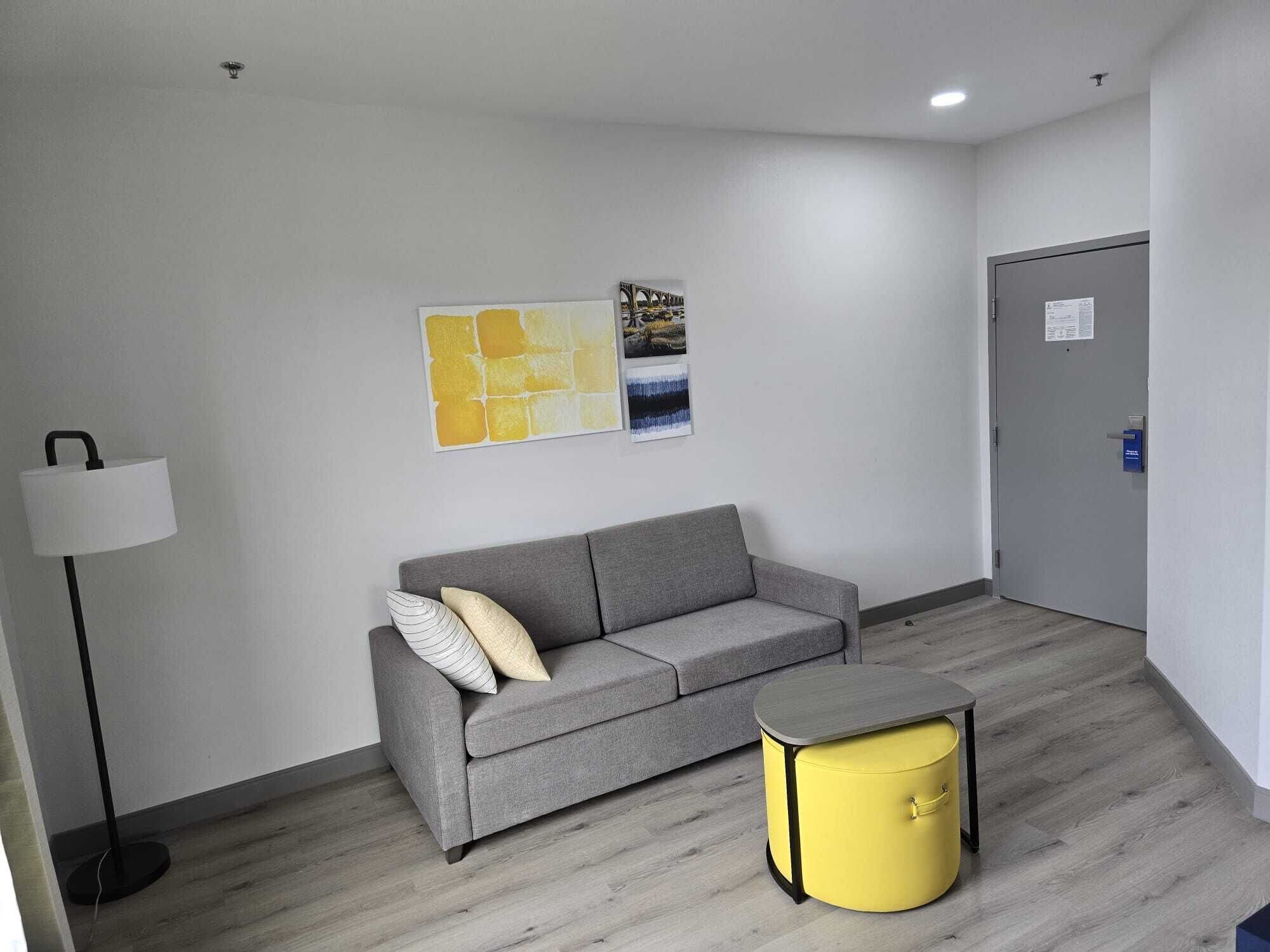 Ein modernes, minimalistisches Zimmer mit einem grauen Sofa, einem gelben Hocker und einer Stehlampe.