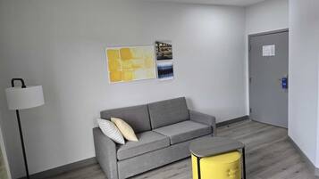 Ein modernes, minimalistisches Zimmer mit einem grauen Sofa, einem gelben Hocker und einer Stehlampe.