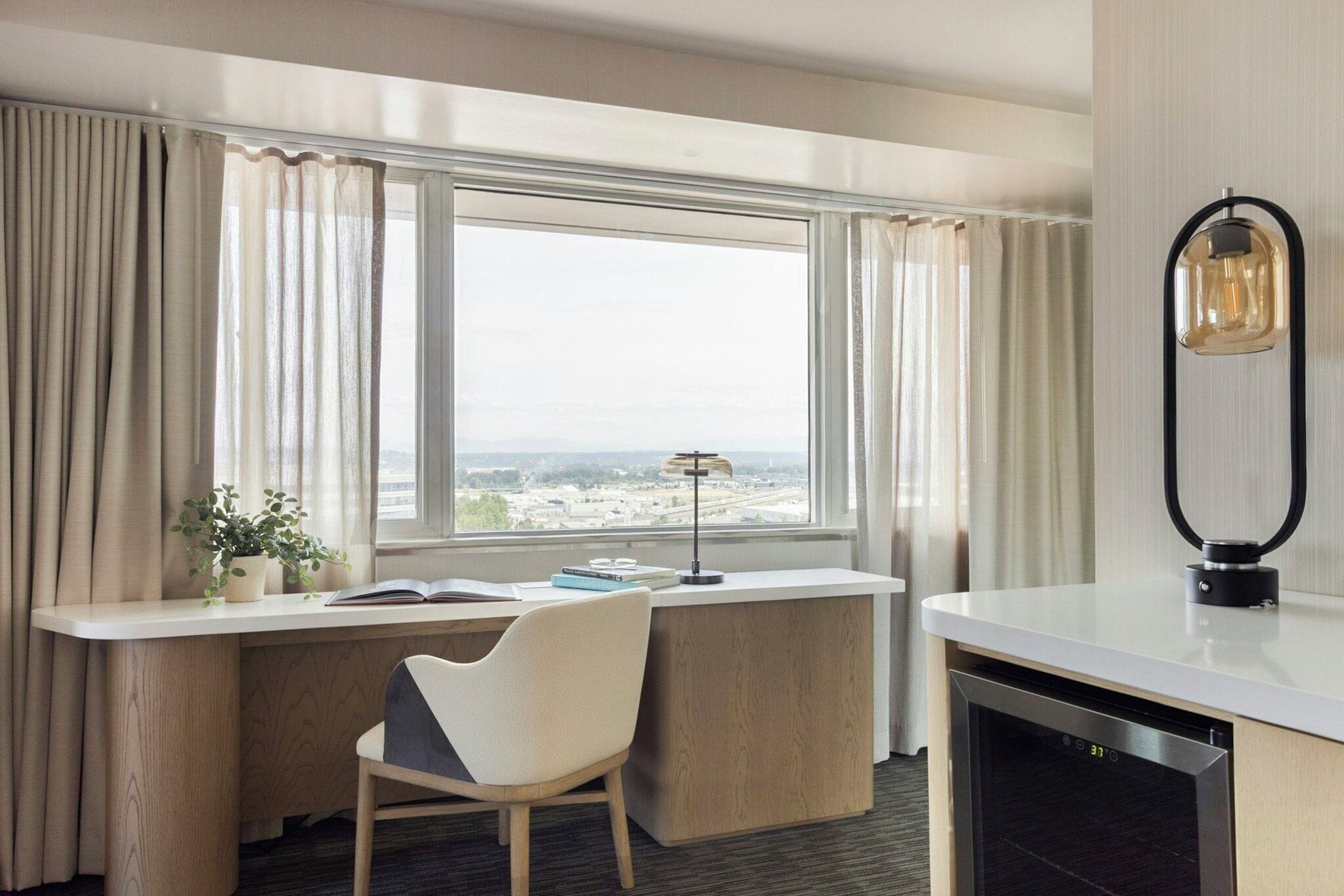 A hotel room with a desk, chair, and a view of a cityscape through large windows.