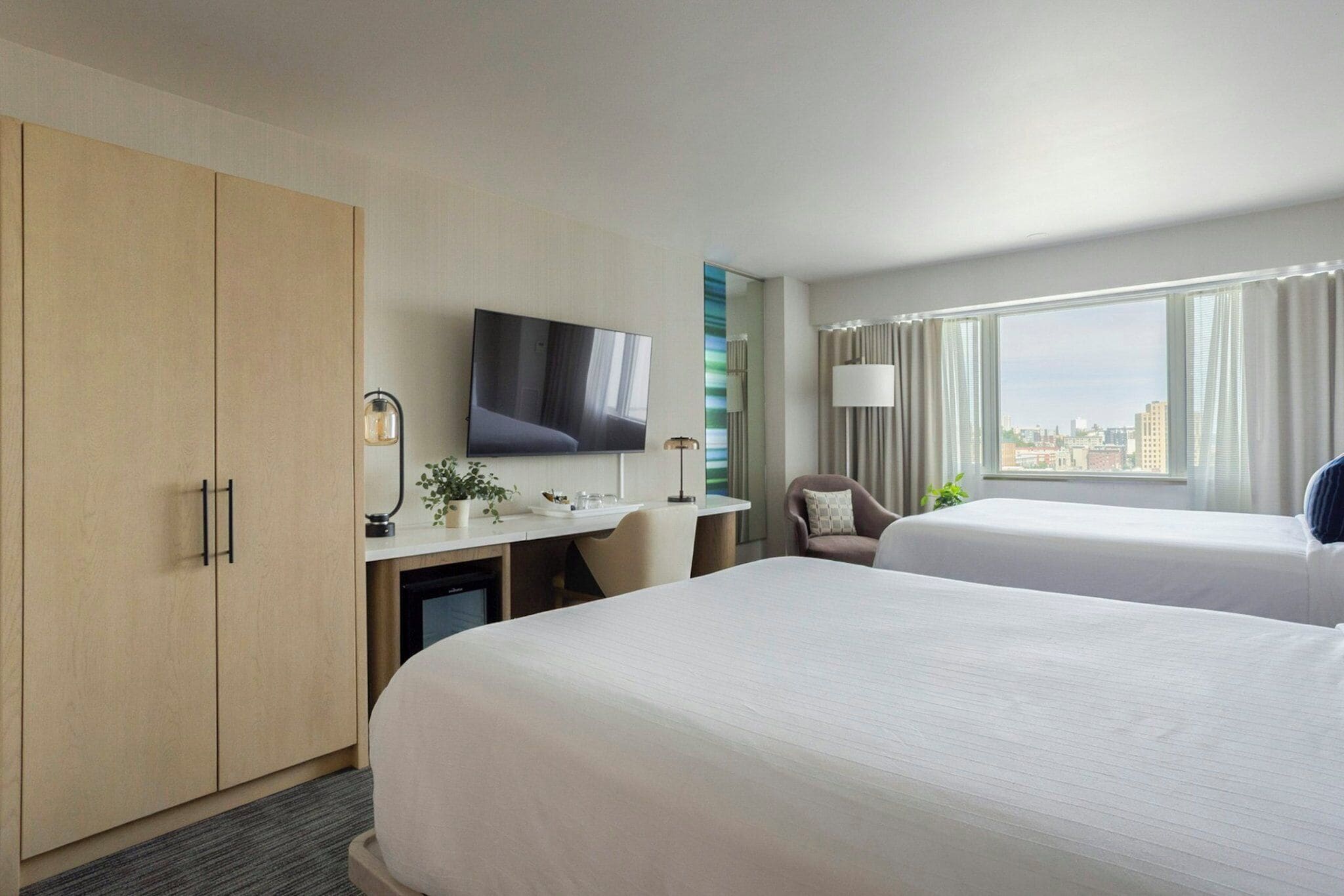 Habitación de hotel con dos camas, un escritorio, un televisor de pantalla plana, una silla y vistas a la ciudad desde la ventana.