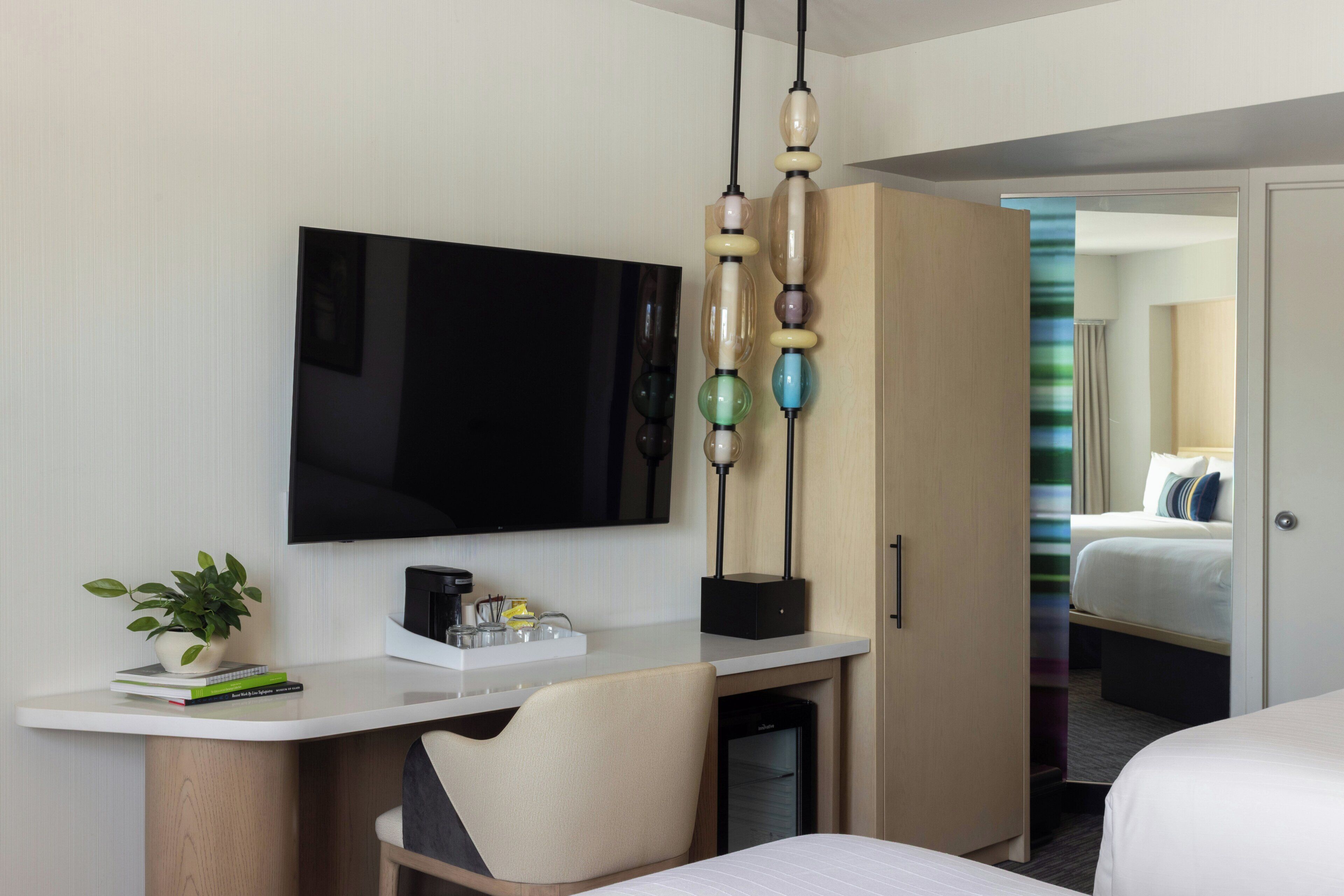 Quarto de hotel com televisão de tela plana, mesa com cafeteira, uma cadeira, guarda-roupa e uma cama.