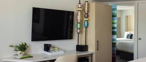 Quarto de hotel com televisão de tela plana, mesa com cafeteira, uma cadeira, guarda-roupa e uma cama.