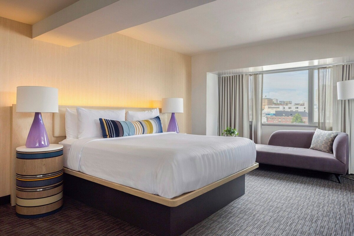 A hotel room with a large bed, two bedside lamps, a sofa, and a view of the city through the window.