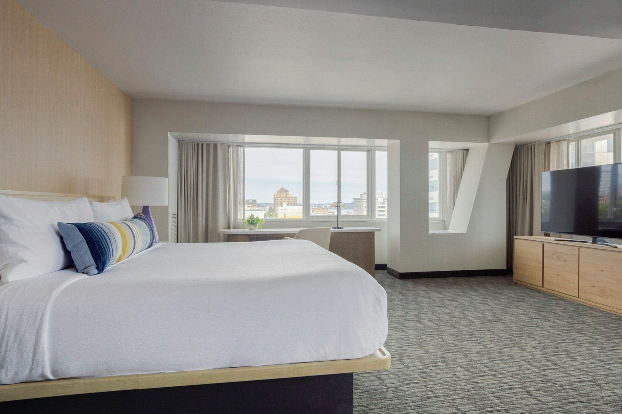 A hotel room with a large bed, a television, a desk, and a window with a city view.