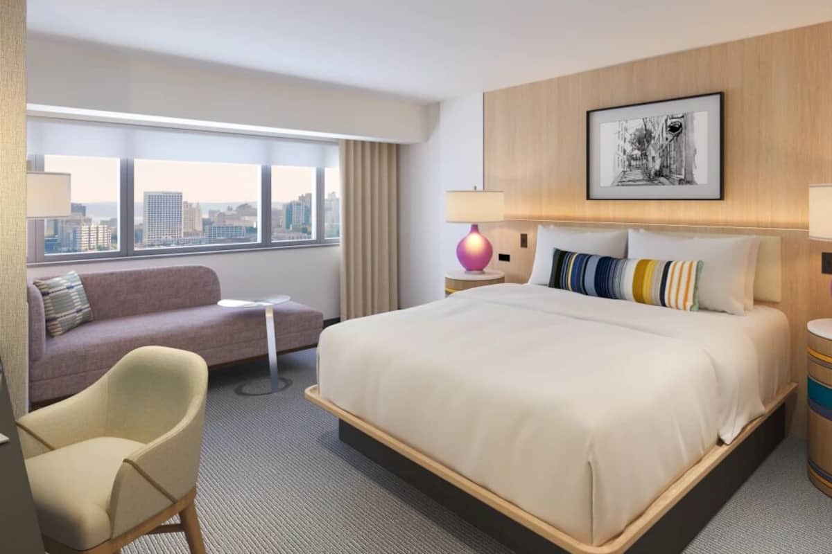 A hotel room with a bed, a sofa, a chair, a desk, a lamp, a picture on the wall, and a view of the city.
