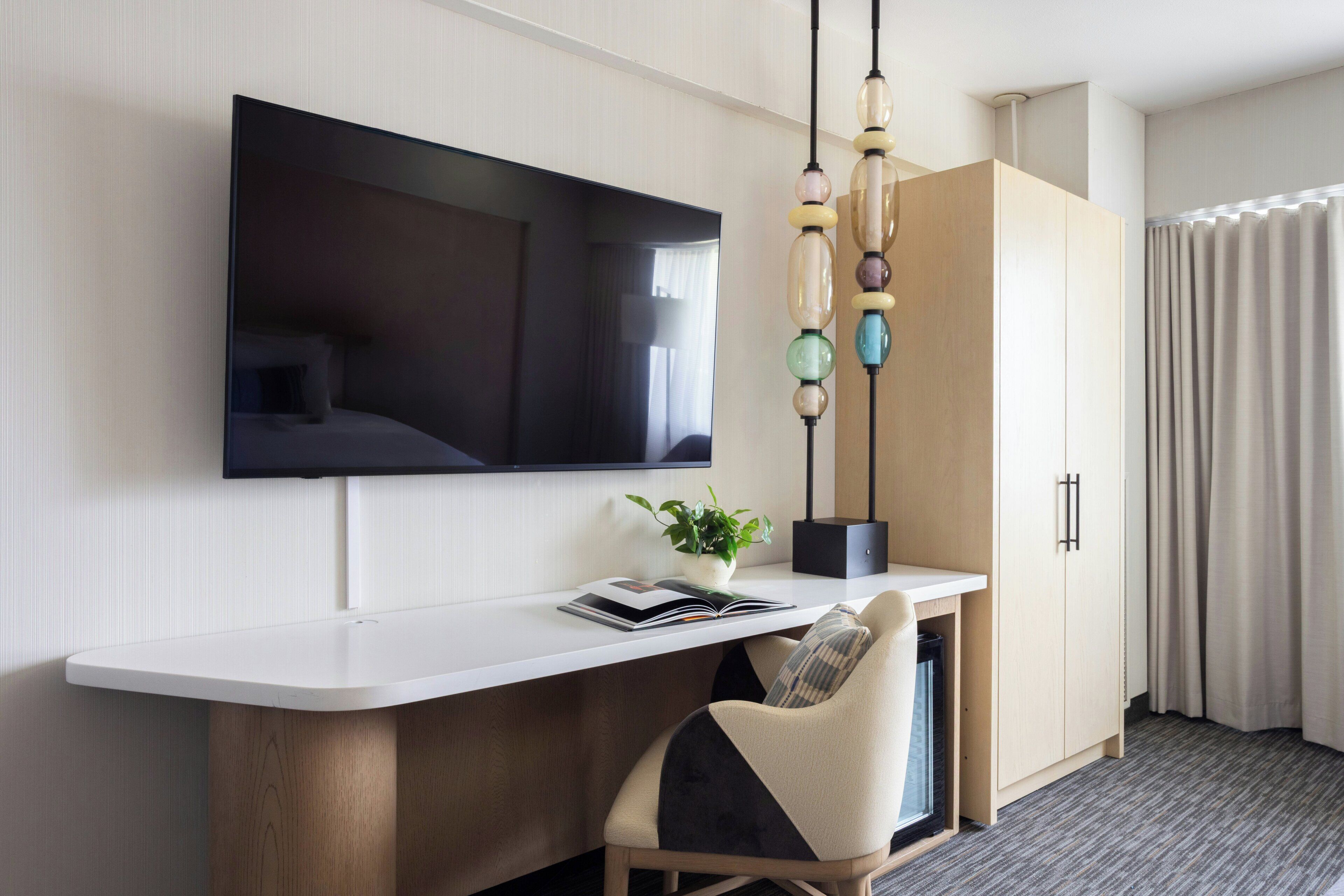 Une chambre d’hôtel équipée d’un téléviseur à écran plat fixé au mur, d’un bureau avec une chaise, d’une plante en pot et d’une armoire.