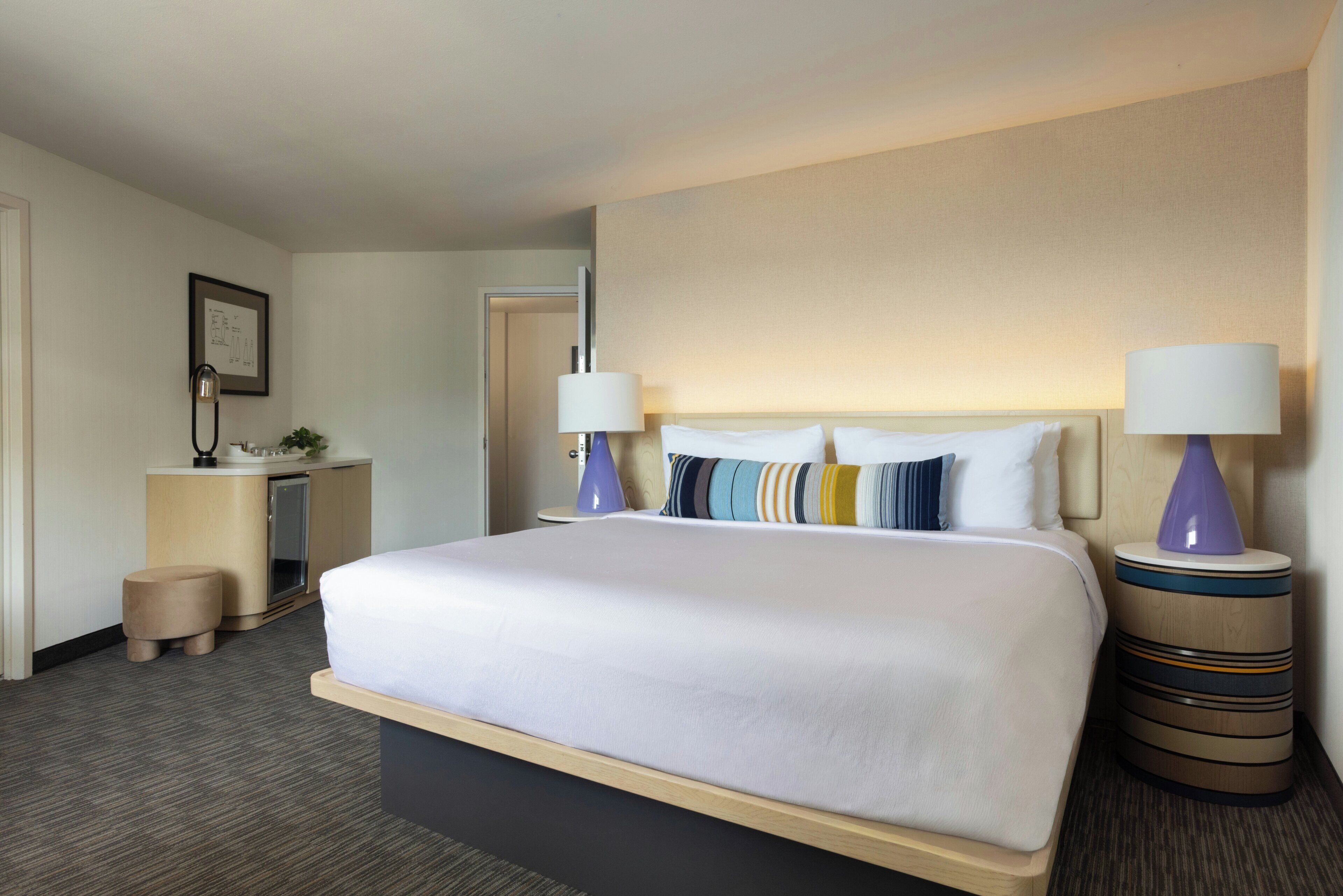 A hotel room with a large bed, two bedside lamps, a small desk with a lamp, a plant, and a framed picture on the wall.