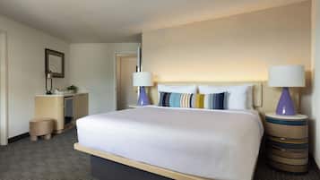 A hotel room with a large bed, two bedside lamps, a small desk with a lamp, a plant, and a framed picture on the wall.