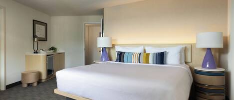 A hotel room with a large bed, two bedside lamps, a small desk with a lamp, a plant, and a framed picture on the wall.