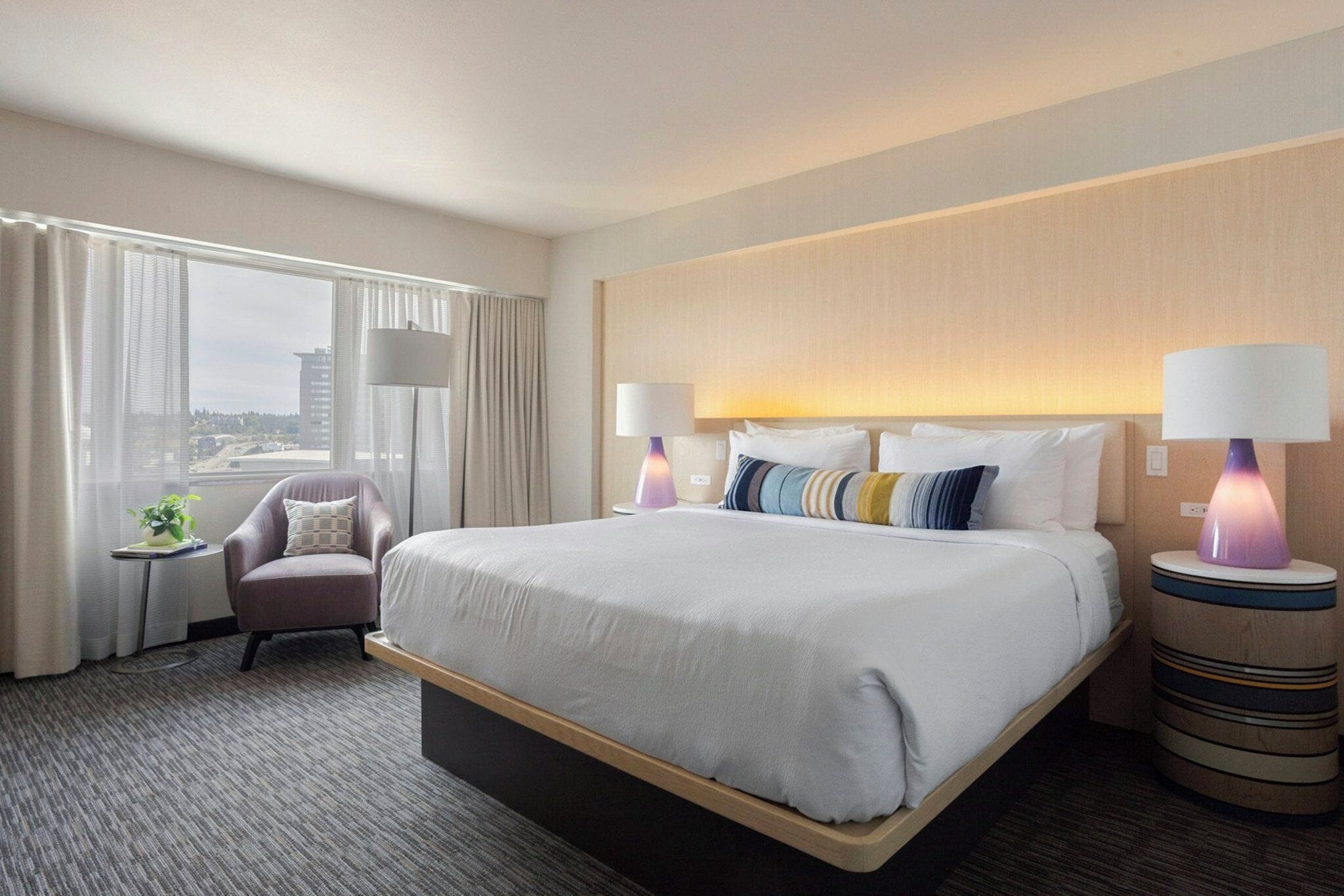 A hotel room with a large bed, two bedside lamps, a chair, a small table, and a view of the city.