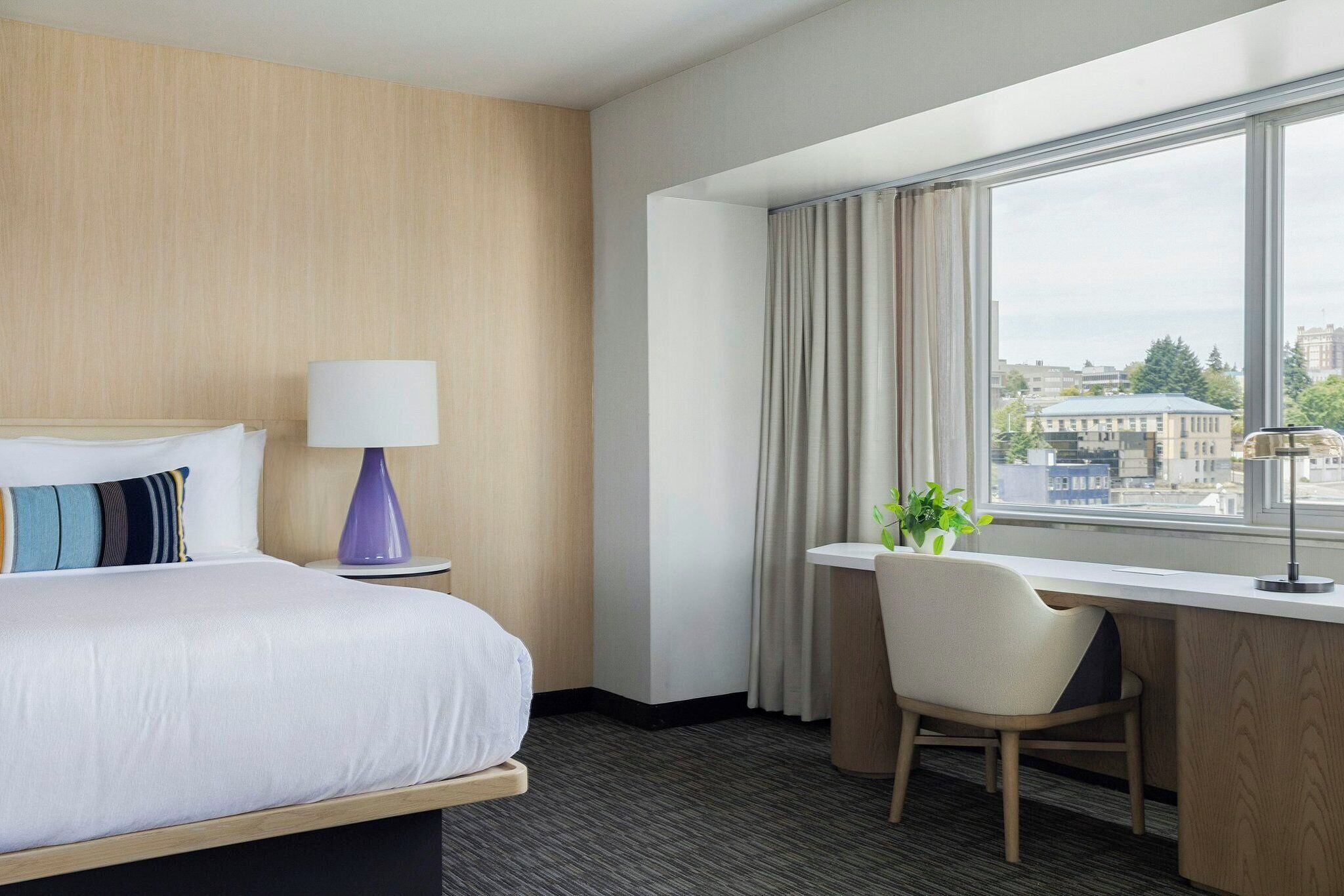 A hotel room with a bed, a desk, a chair, a lamp, and a window with curtains.