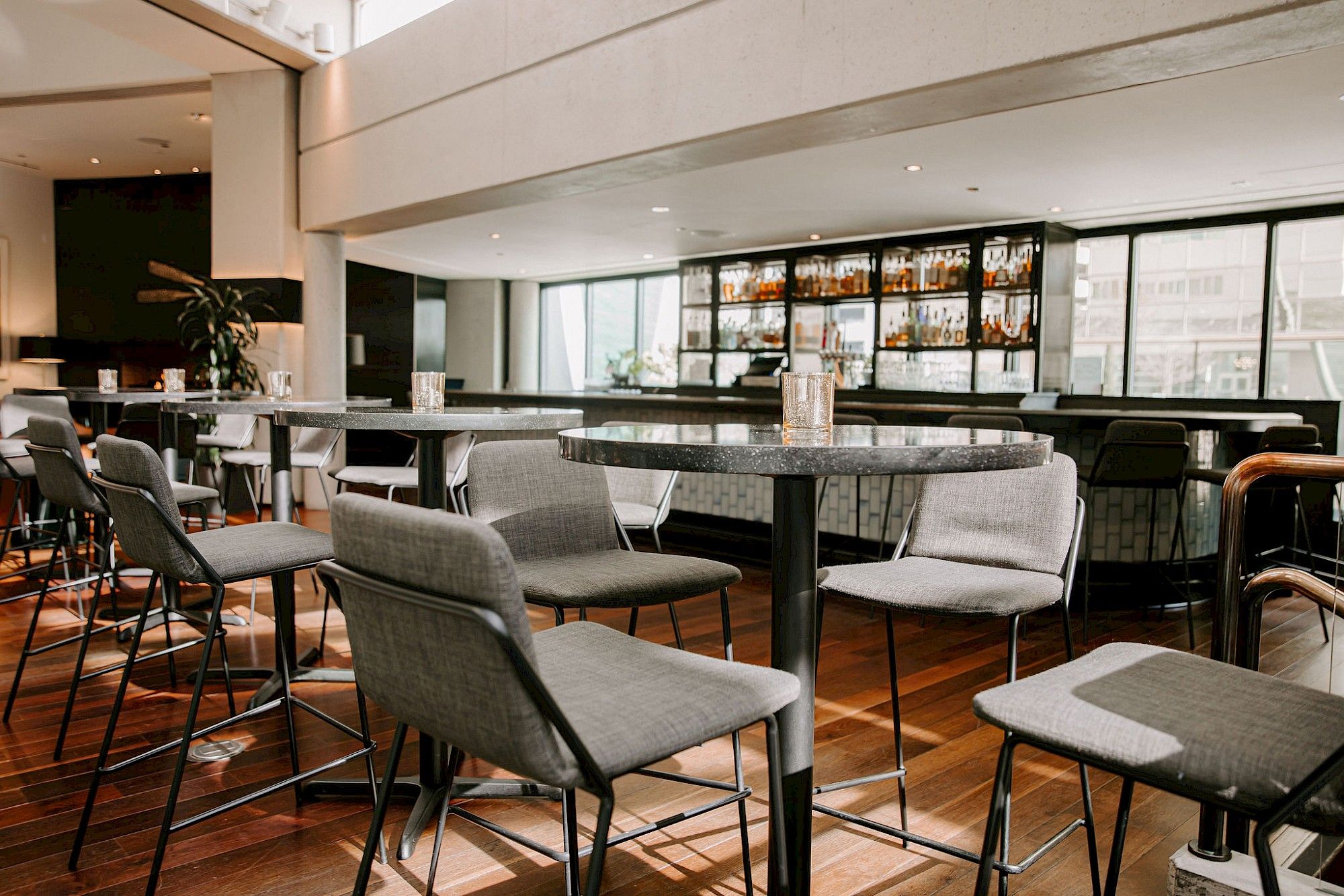 A modern bar area with round tables and grey upholstered chairs, featuring a large window with a view of the city.