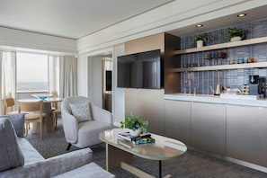 Habitación de hotel moderna con zona de estar, una mesa de centro de vidrio, un televisor de pantalla plana, una kitchenette con salpicadero de azulejos y vistas a la ciudad.