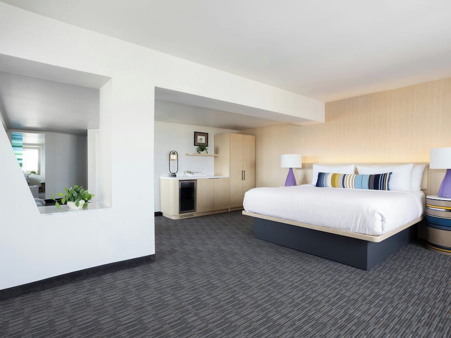 A modern hotel room with a large bed, bedside lamps, a small desk, and a view of the adjacent room.