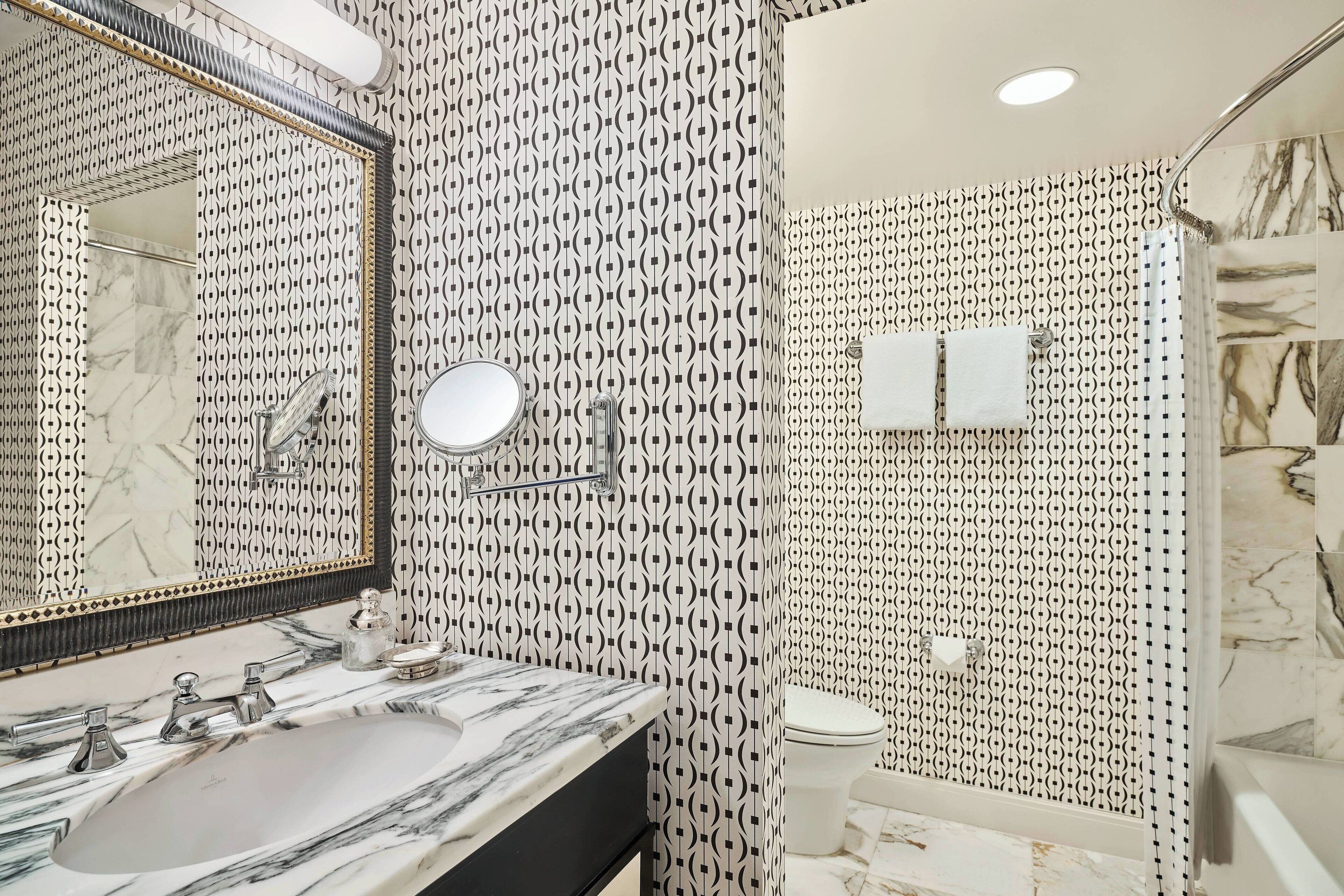 A bathroom with a patterned wall, a large mirror, a sink with a marble countertop, a toilet, and a shower area.