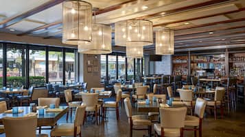 A spacious restaurant with multiple tables and chairs, large windows, and hanging light fixtures.