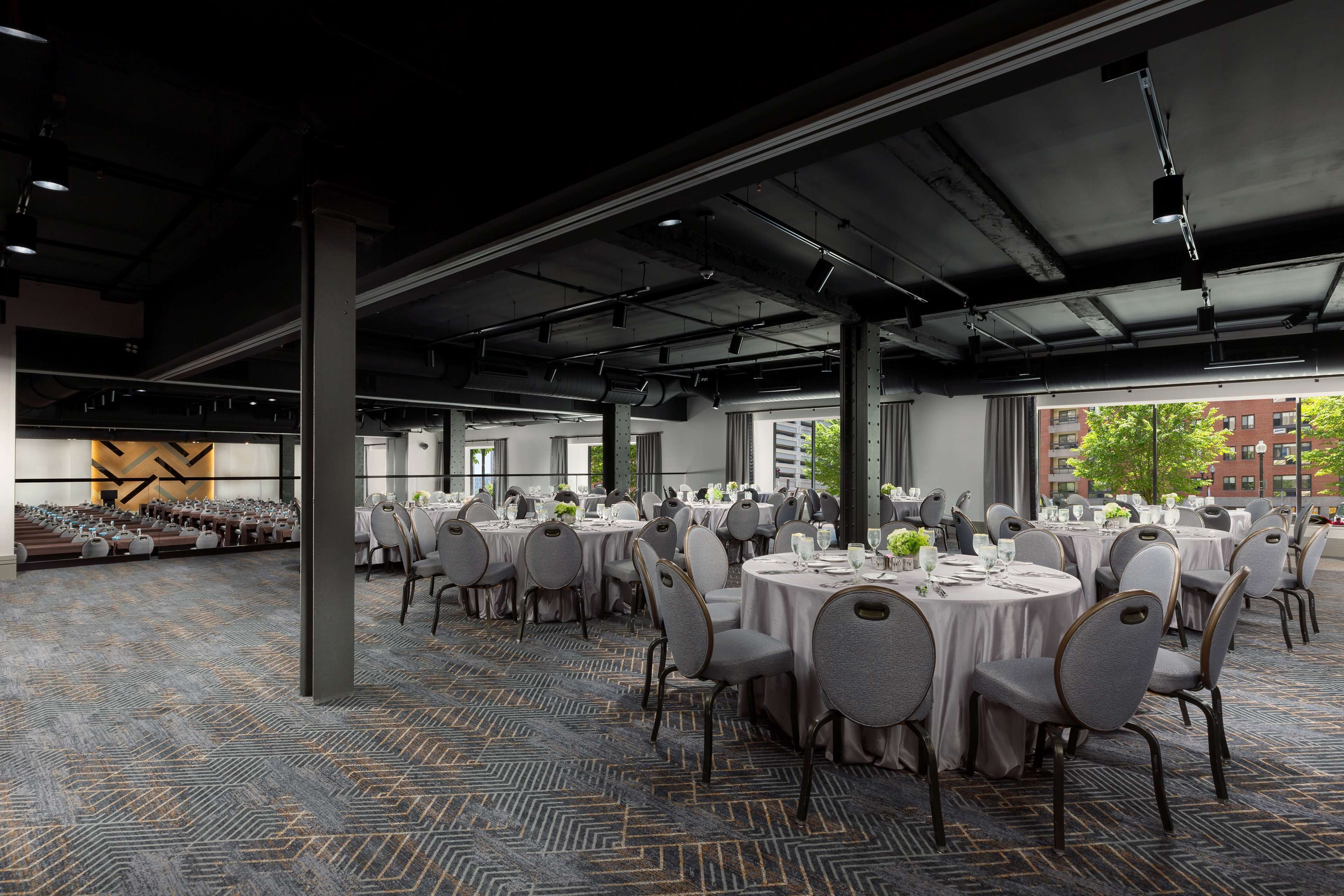 A spacious banquet hall with round tables and chairs, large windows, and a patterned carpet.