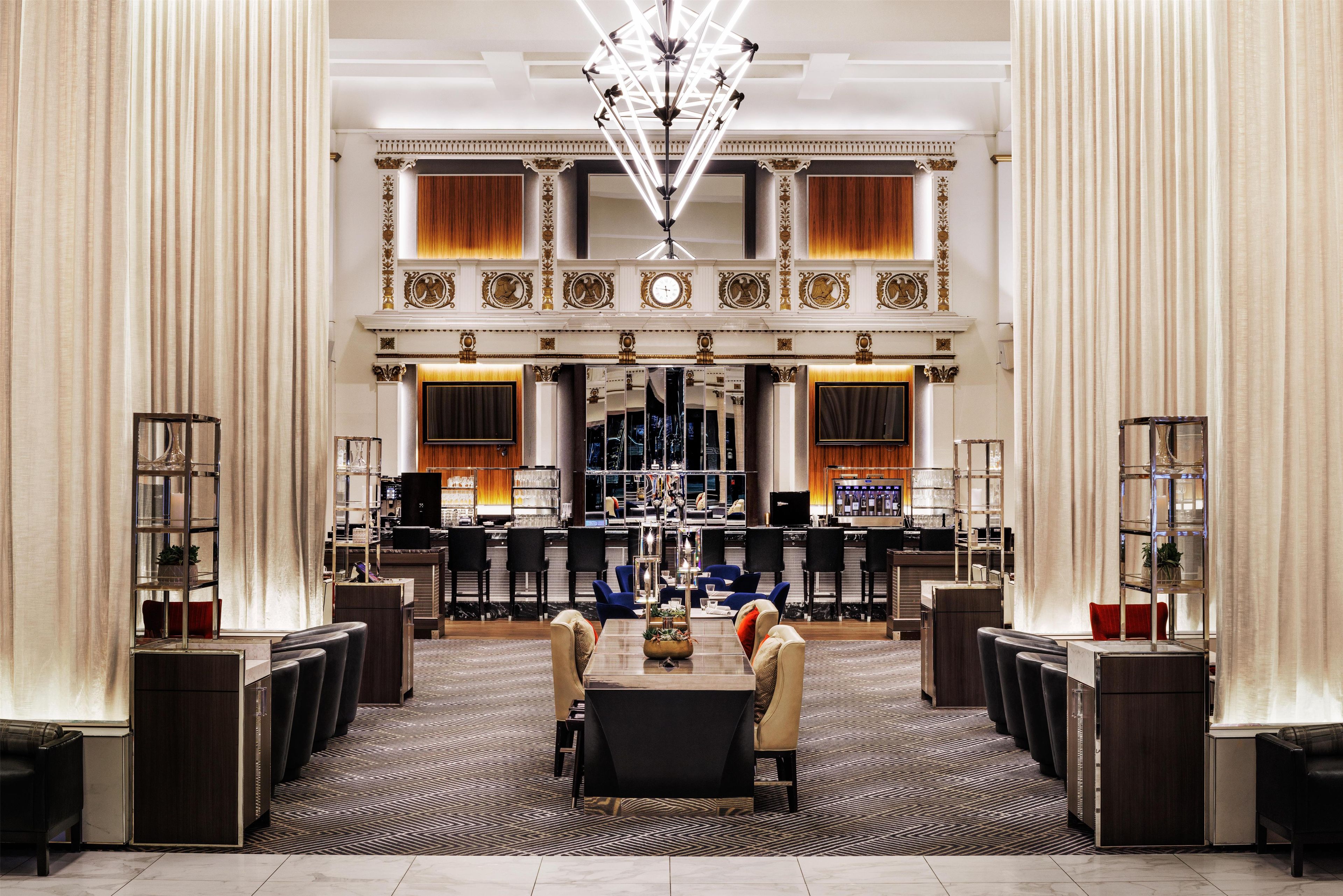 Un hall espacioso con techo alto, columnas ornamentales, una lámpara central y una zona de bar con asientos.