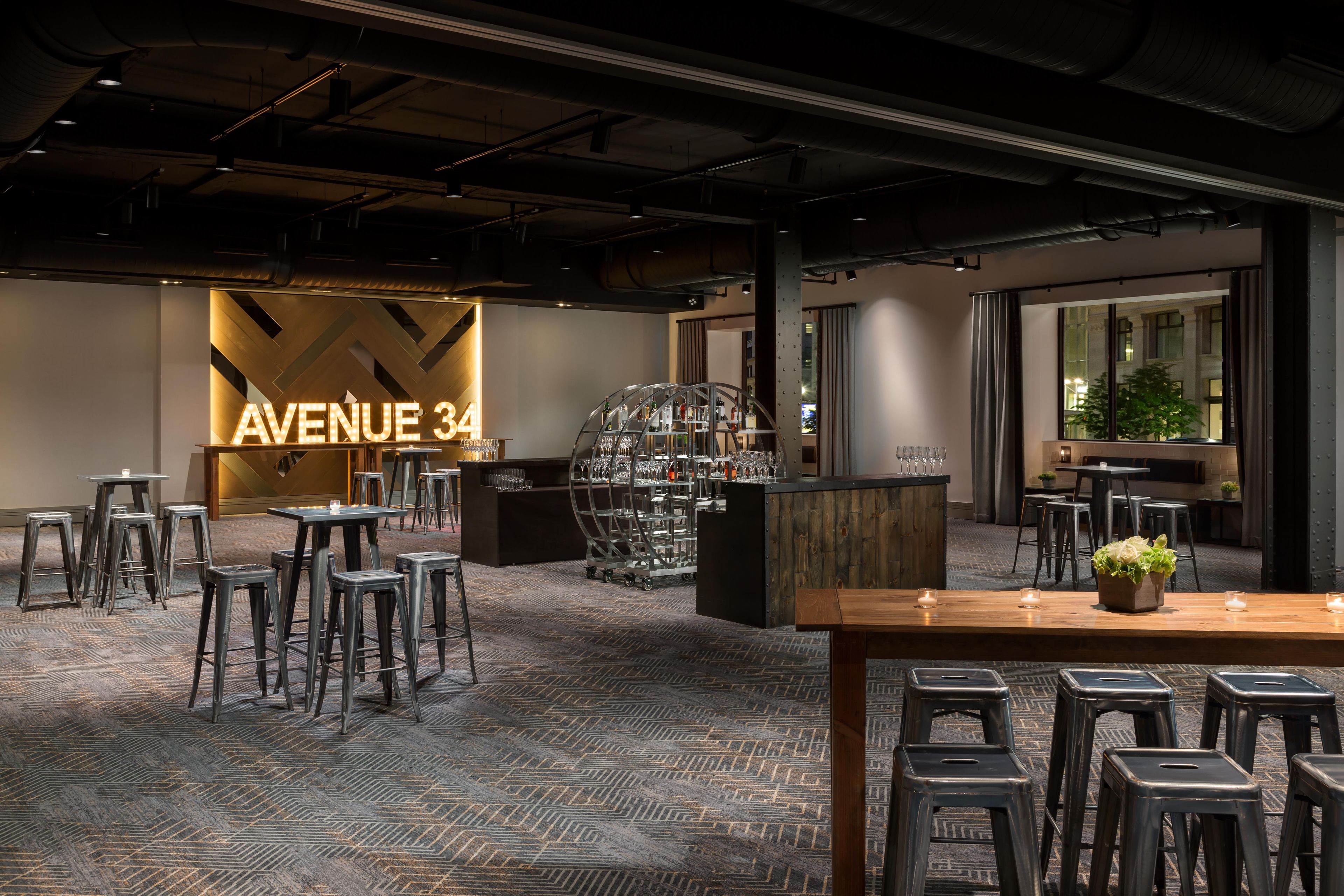 Een moderne bar met een opvallend 'AVENUE 34' bord, barkrukken, een toonbank met glazen en een kamerbreed tapijt met dessin.