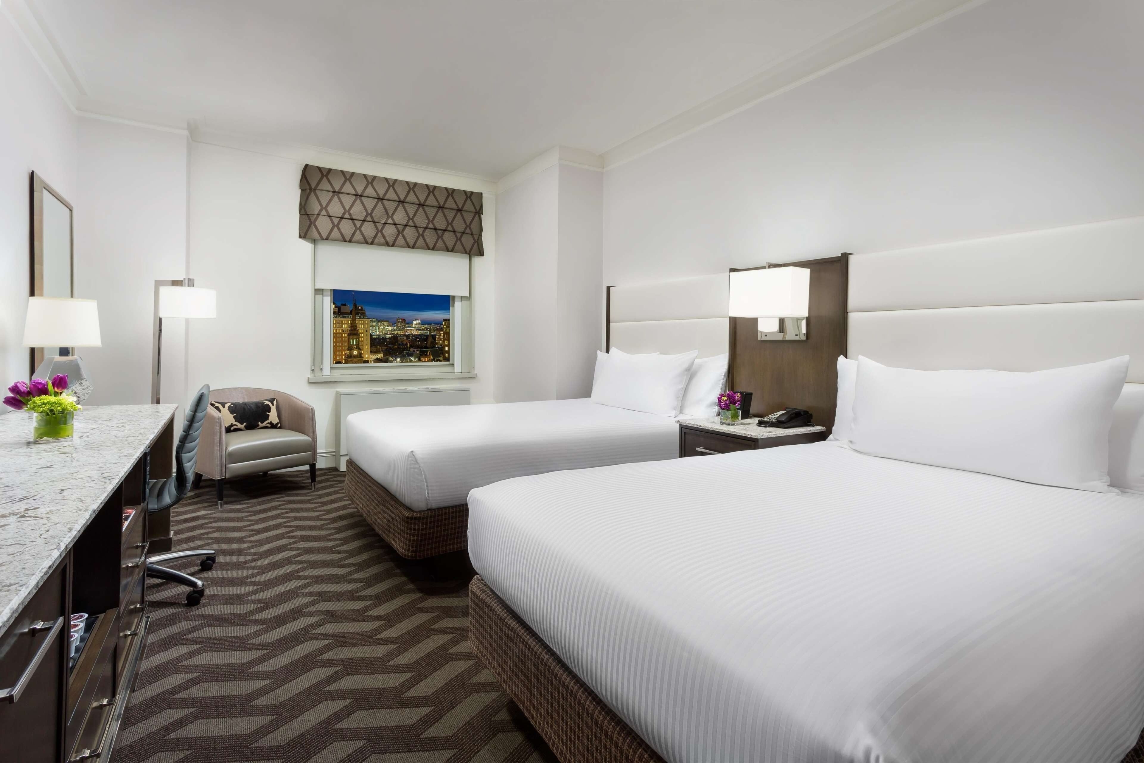 A hotel room with two beds, a desk with a chair, a window with a city view, and a patterned carpet.