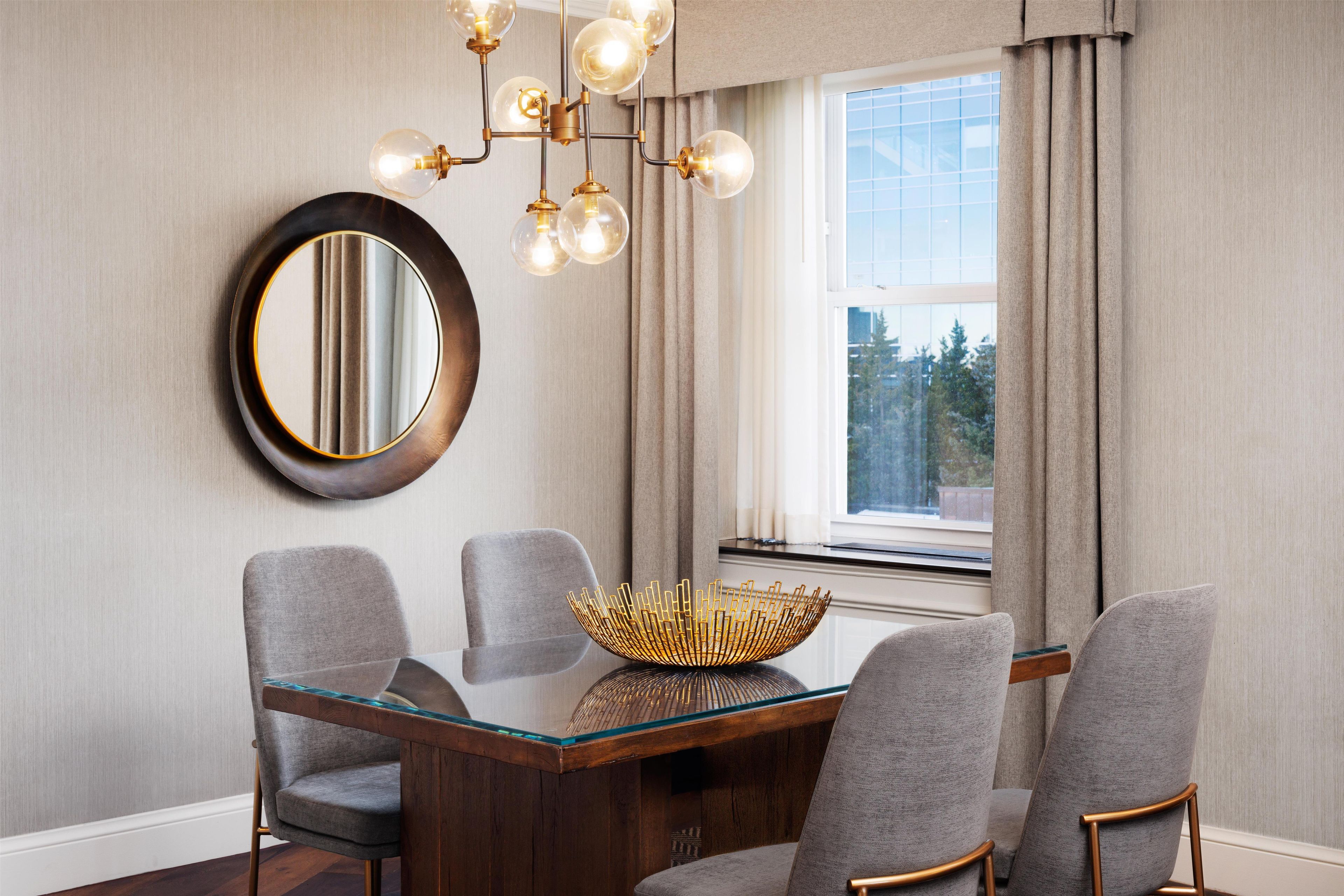 A dining area with a round table, four chairs, a chandelier, a mirror, and a window with curtains.