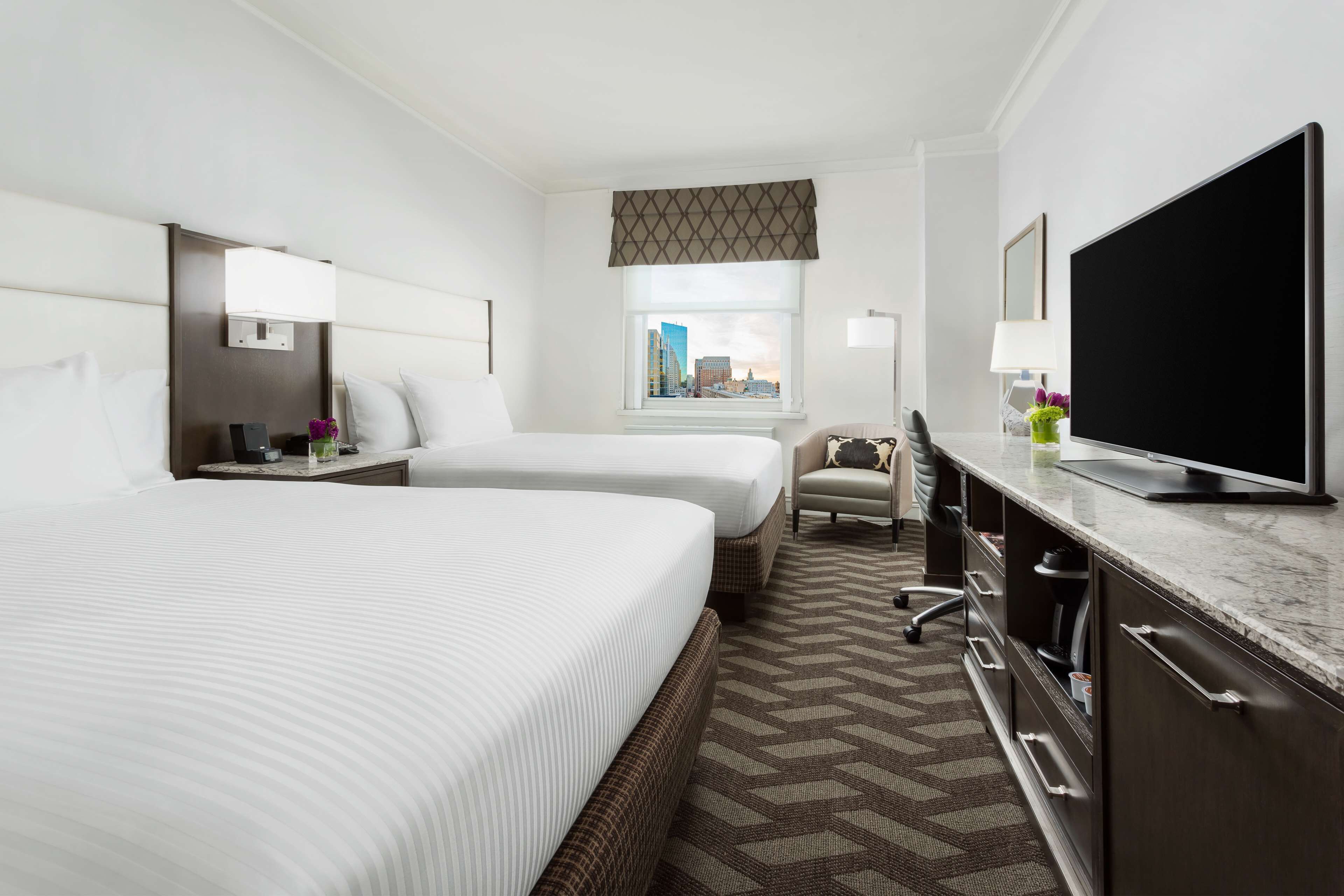 Habitación de hotel con dos camas, un escritorio, un televisor de pantalla plana, una ventana con vista a la ciudad y una alfombra estampada.