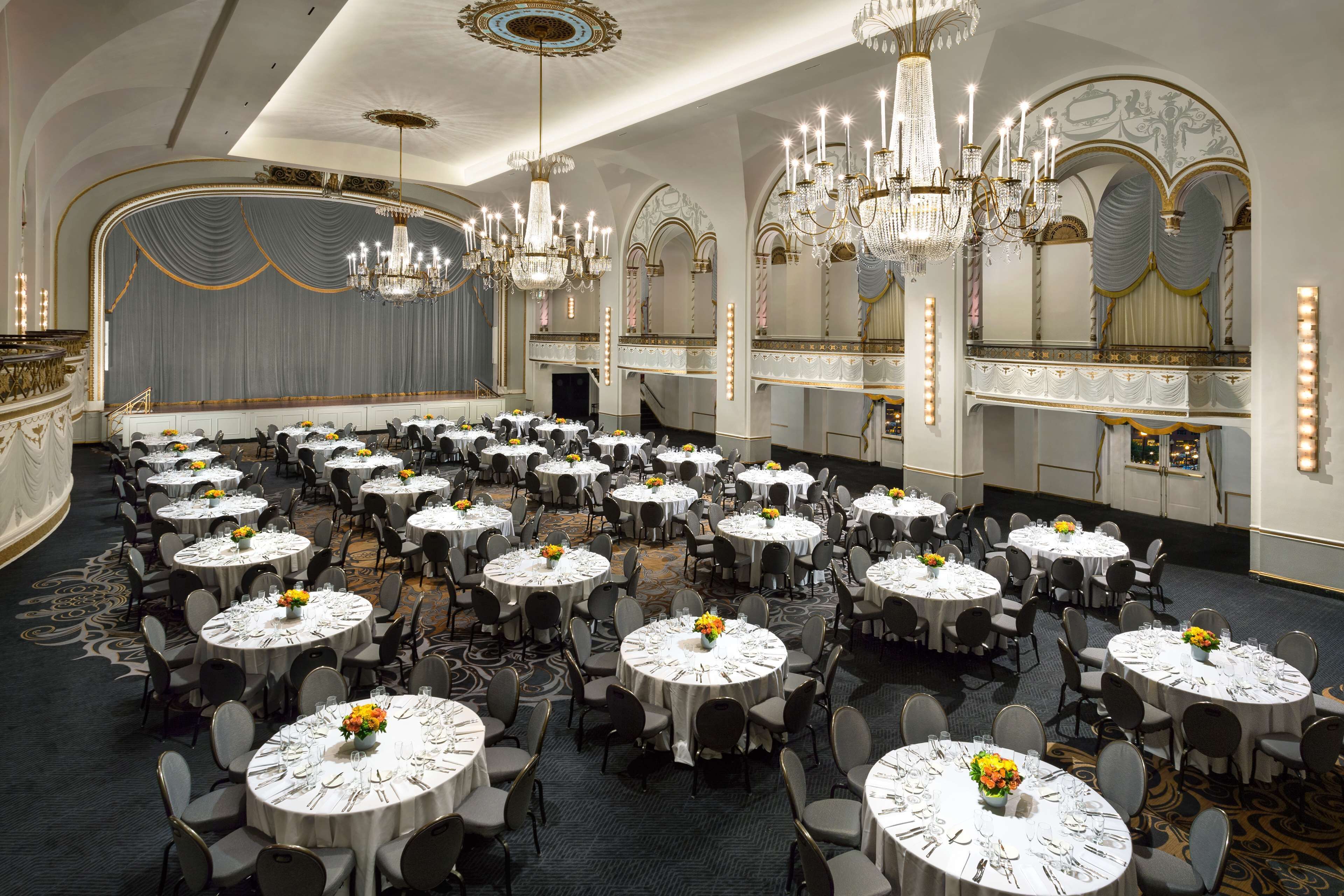 A large banquet hall with round tables, chairs, chandeliers, and a stage.