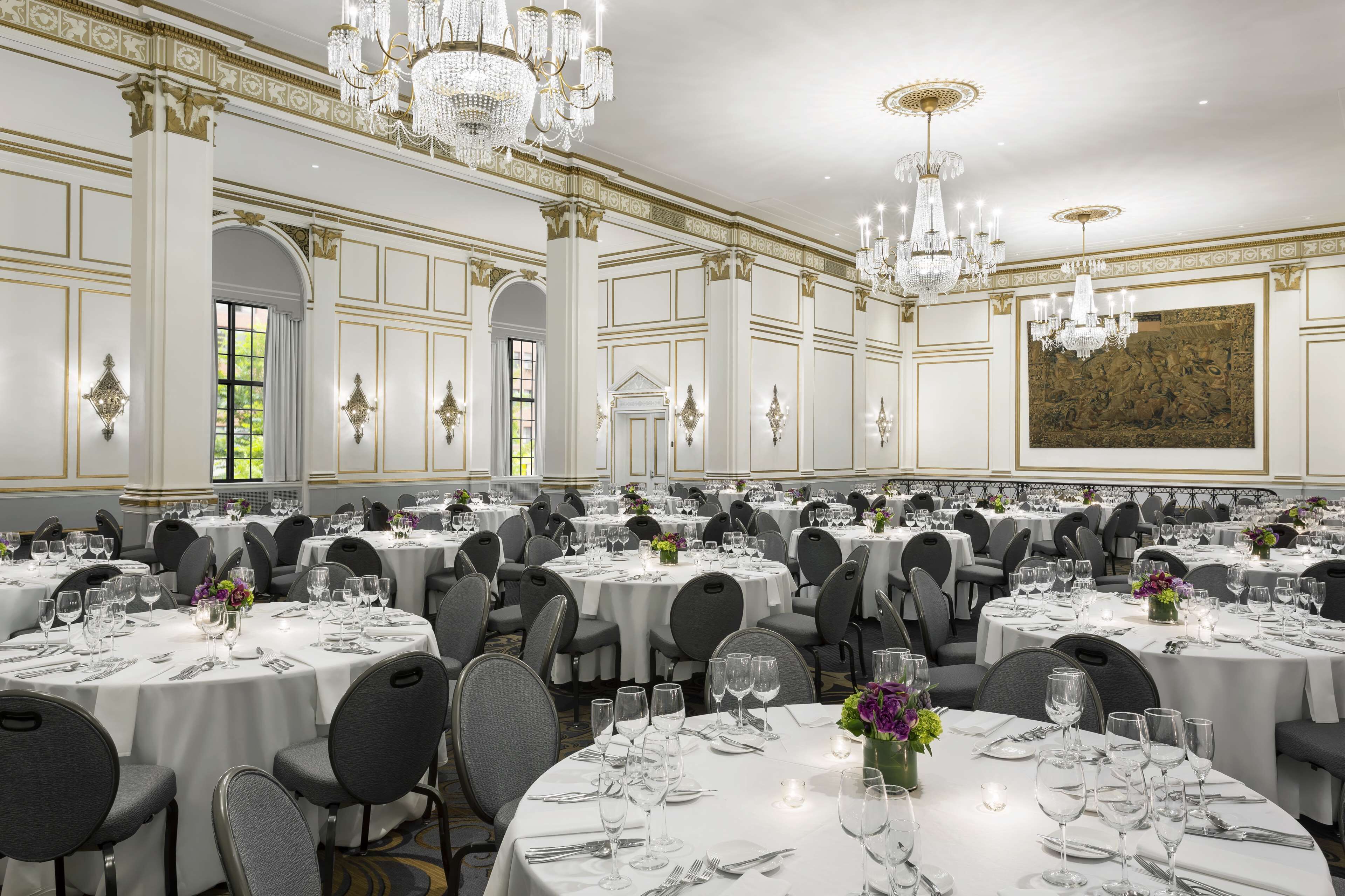 Une salle de banquet avec des tables rondes dressées pour le repas, des lustres et une architecture de style classique.