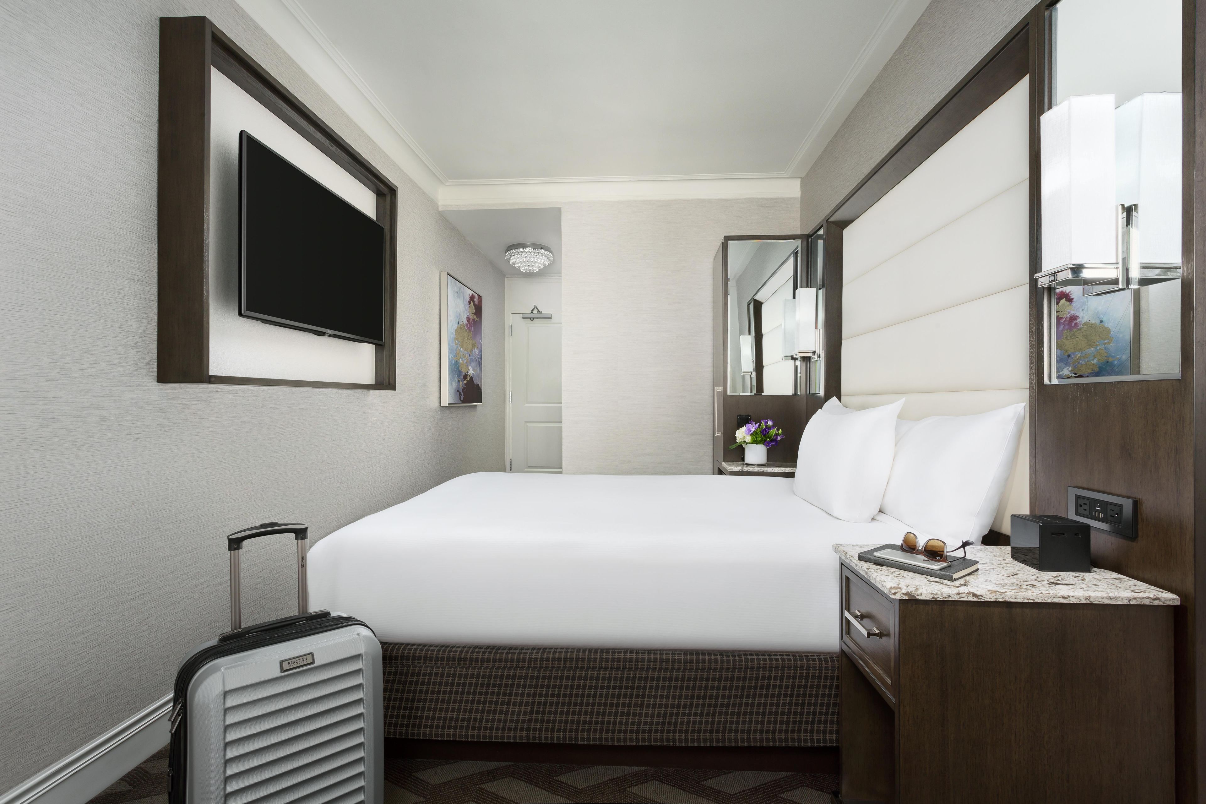 A hotel room with a bed, a flat-screen TV, a bedside table with a lamp, a suitcase, and a mirror.