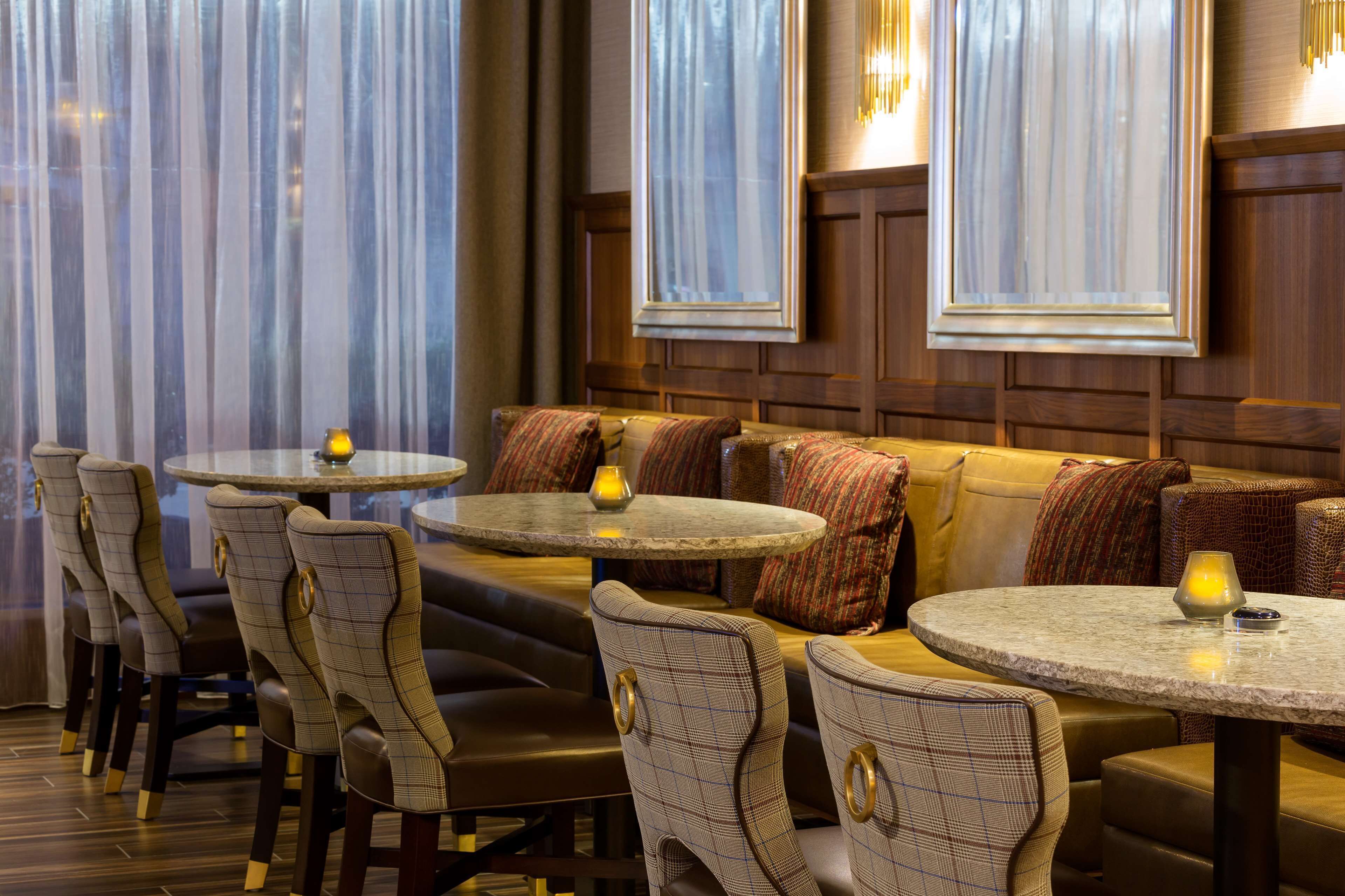 Restaurante com mesas redondas e cadeiras de encosto alto, apresentando uma parede de painéis de madeira e grandes janelas com cortinas leves.