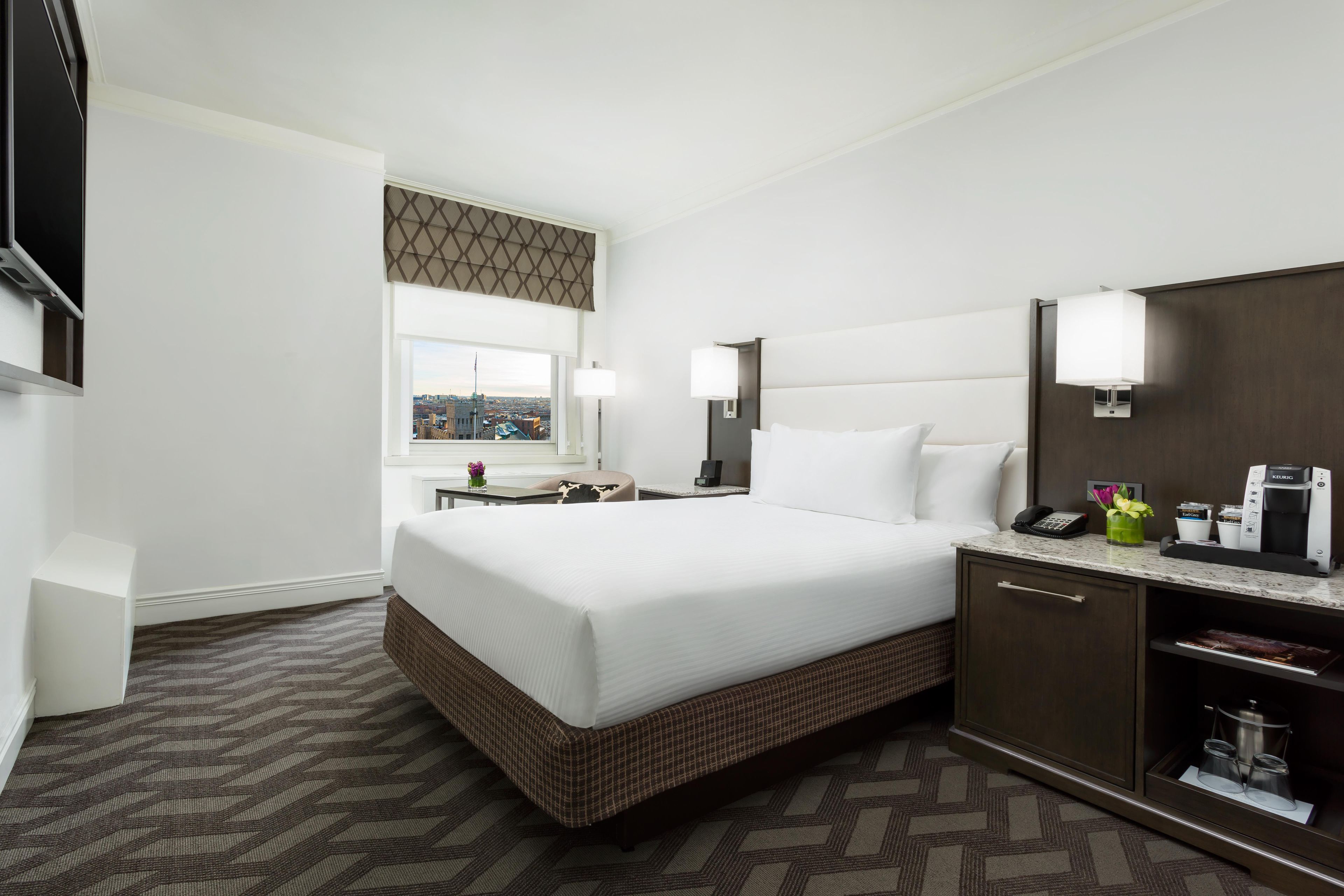 A hotel room with a large bed, a desk with a chair, a minibar, and a view of the cityscape.