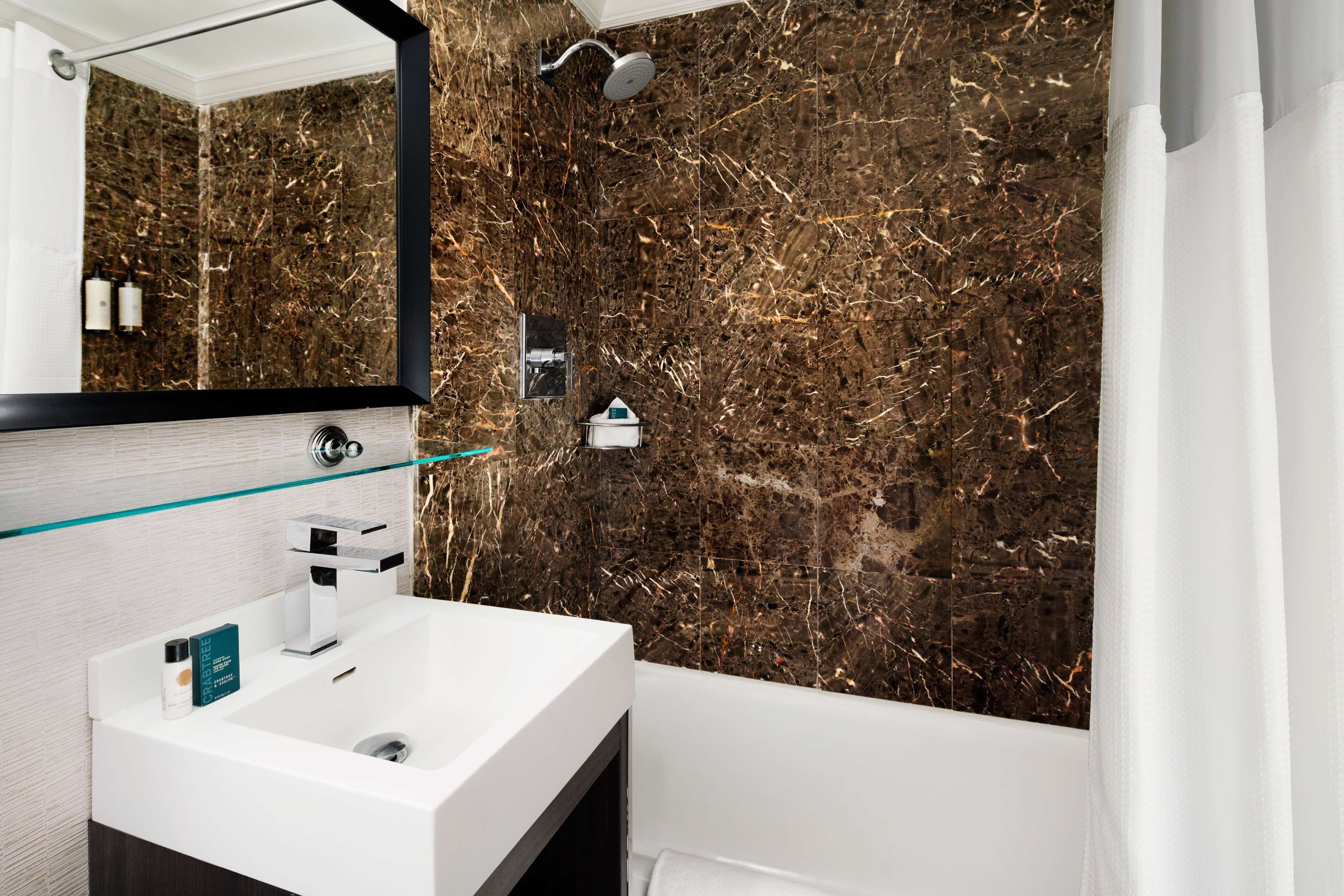 A modern bathroom with a white sink, a glass shelf, a large mirror, and a shower with a marble wall.