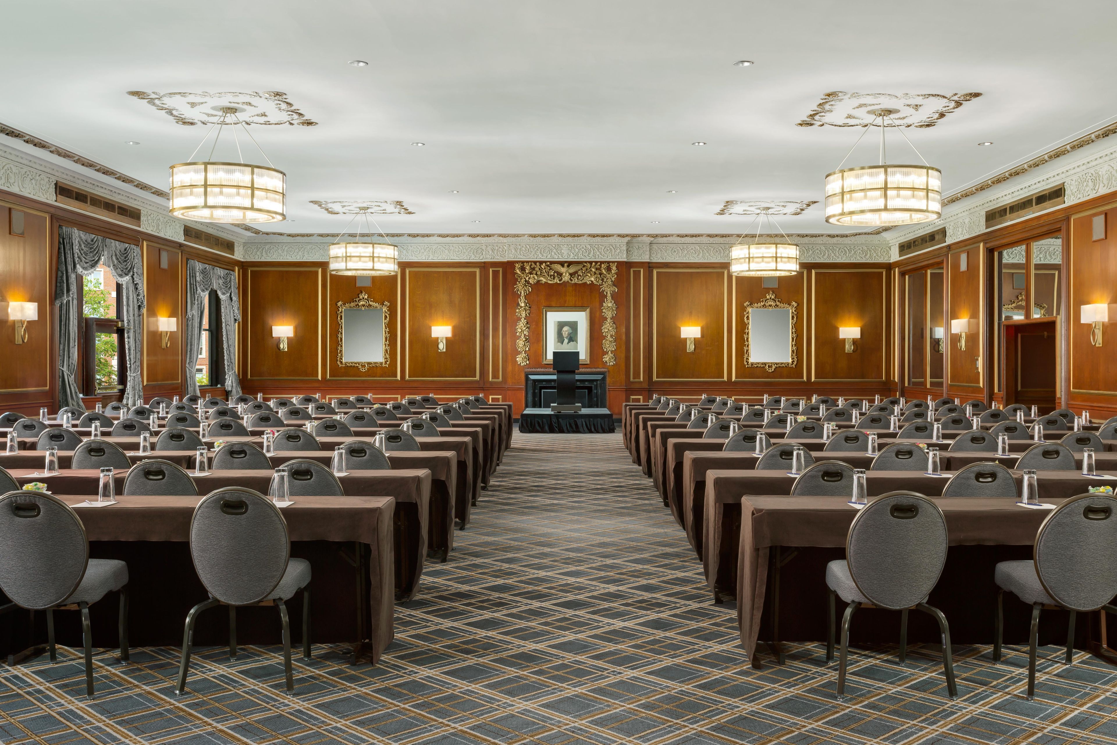 A conference room with rows of tables and chairs, ornate ceiling details, and wall-mounted lamps.