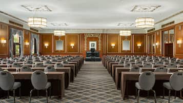 A conference room with rows of tables and chairs, ornate ceiling details, and wall-mounted lamps.