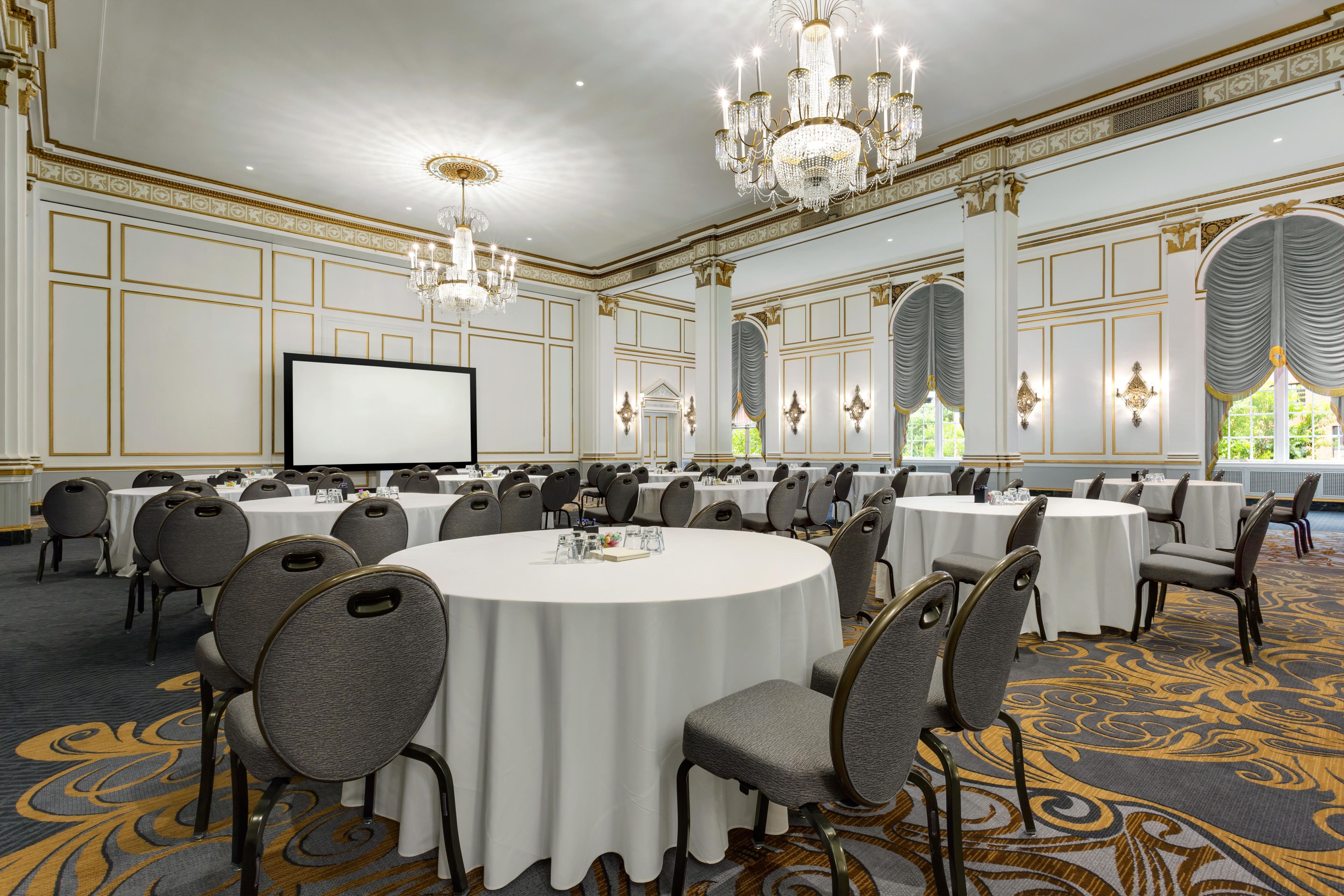 A conference room with round tables, grey upholstered chairs, a large chandelier, and a white screen.
