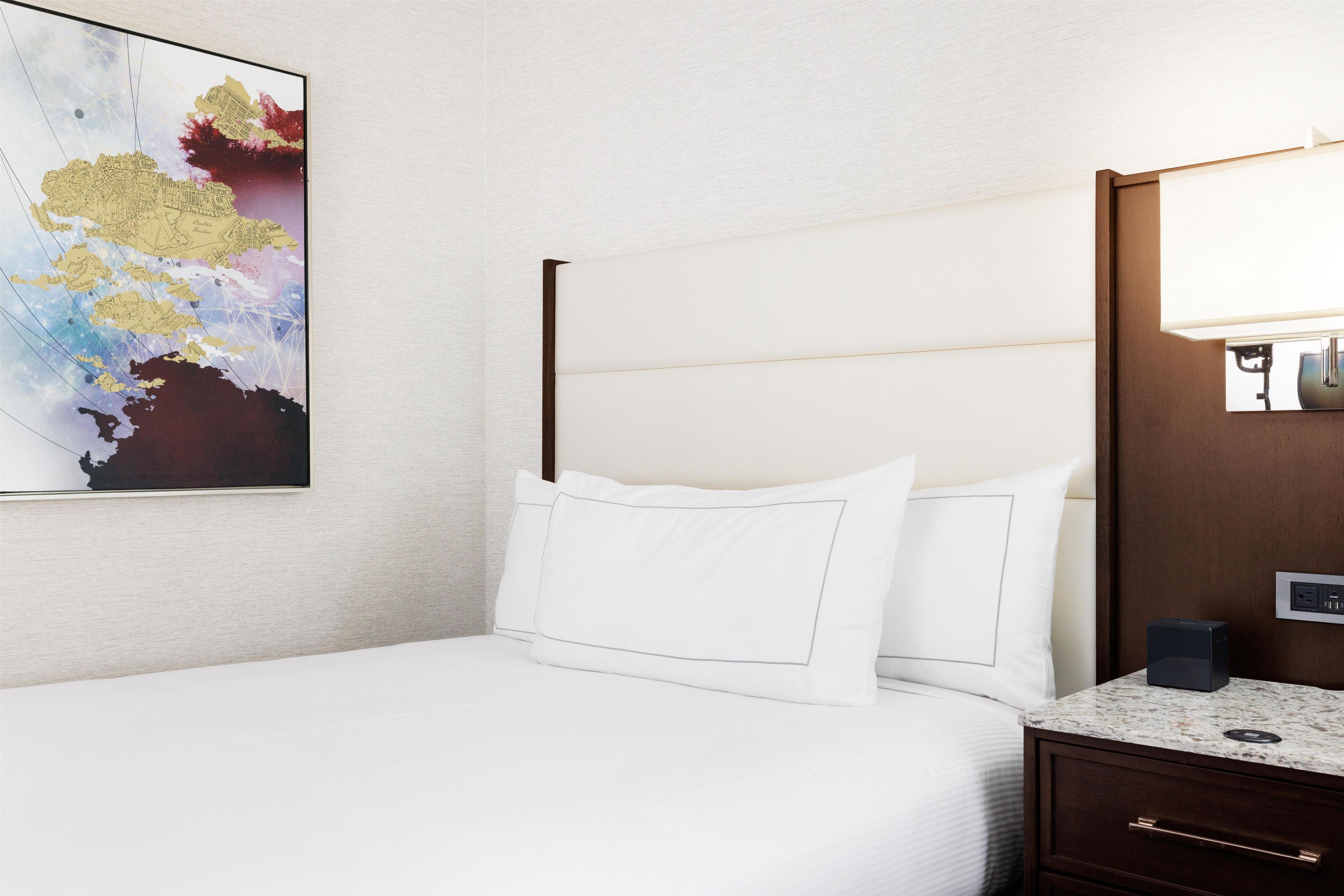 A hotel room with a bed, a nightstand, a lamp, and a wall-mounted artwork.