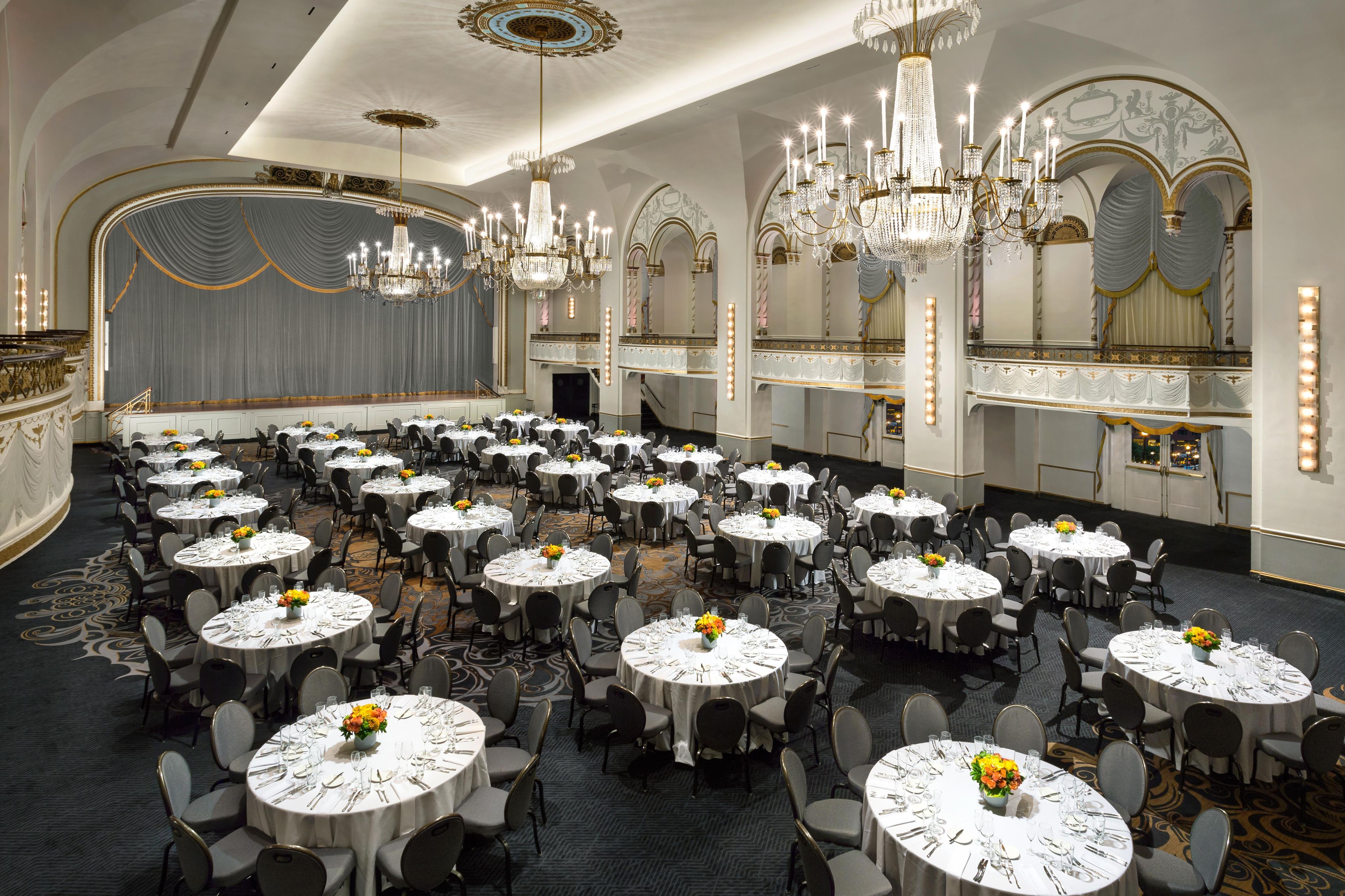 A banquet hall with round tables, chairs, chandeliers, and a stage.