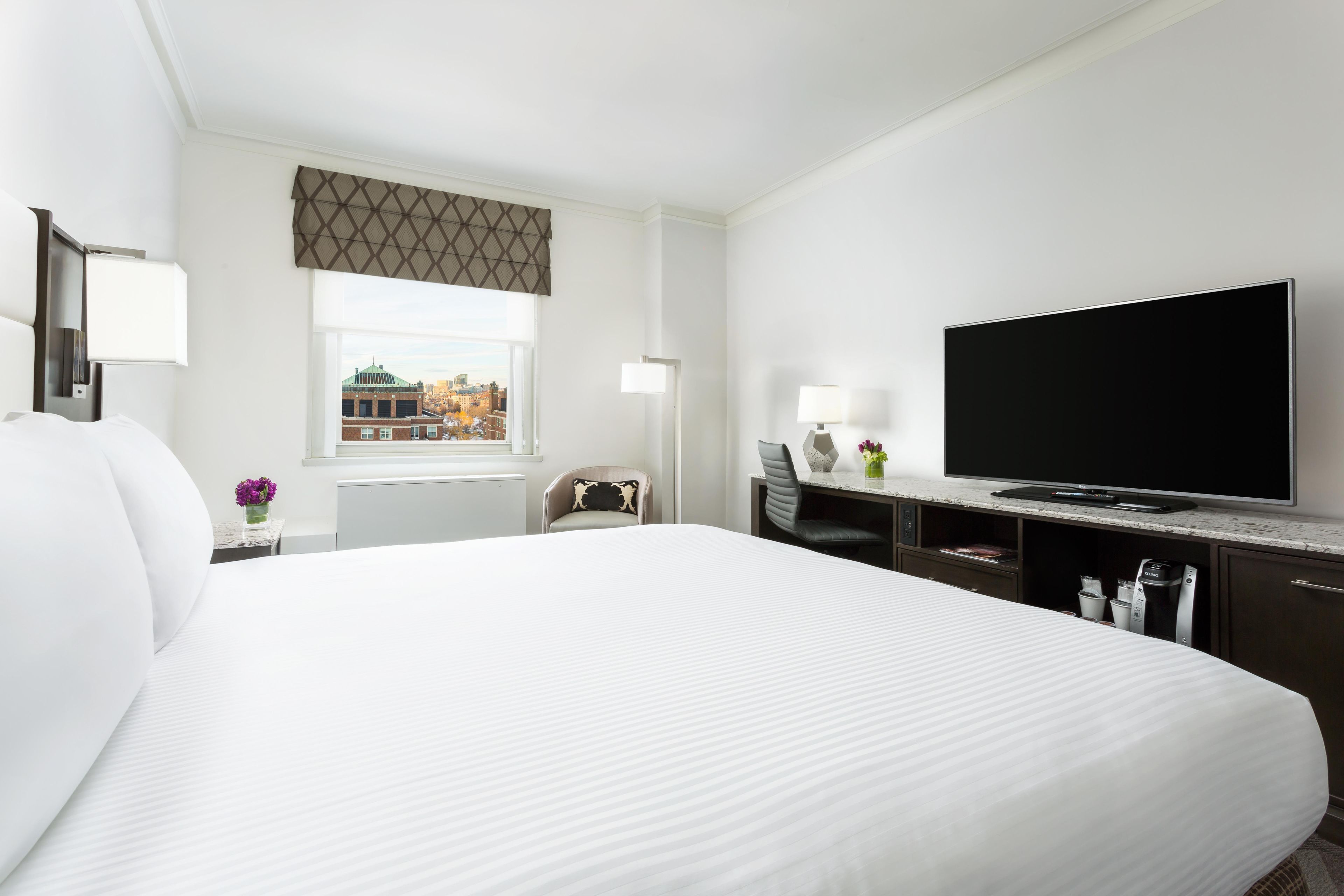 A hotel room with a large bed, a television, a desk with a lamp, and a window with a view of buildings.