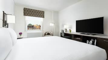 A hotel room with a large bed, a television, a desk with a lamp, and a window with a view of buildings.