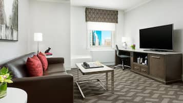 A modern hotel room with a living area, a flat-screen TV, a desk, and a view of the cityscape.