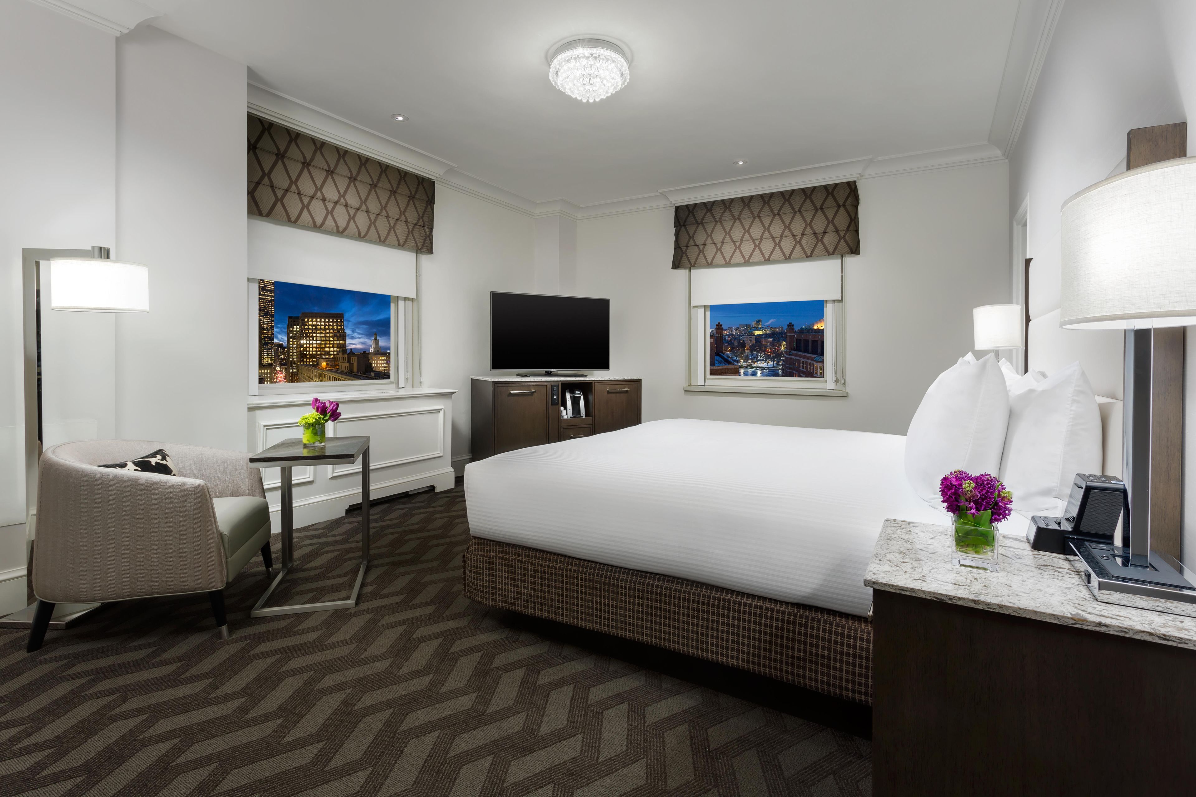 A hotel room with a bed, a desk, a chair, a television, and two windows with city views.