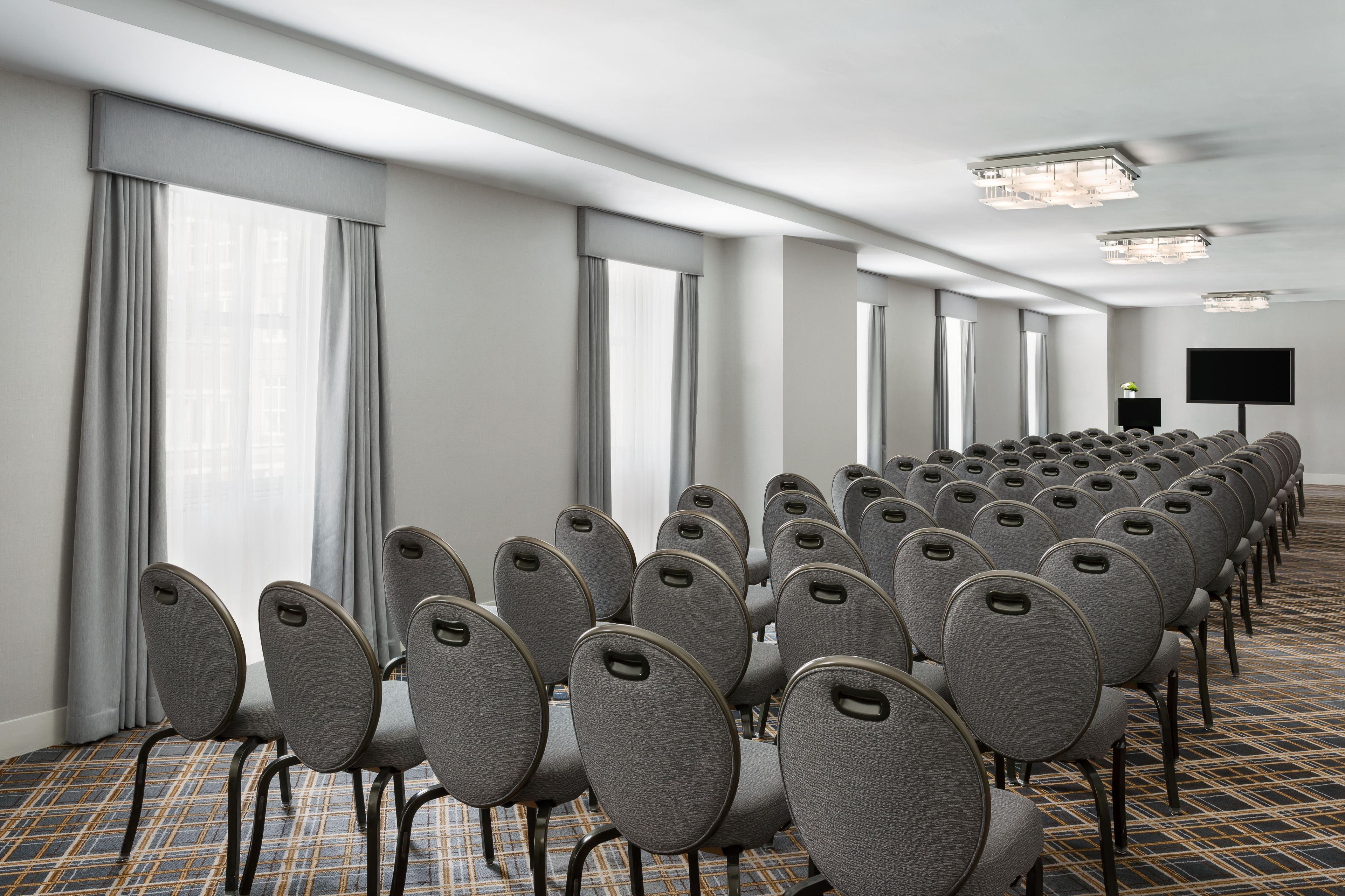 Un salón de conferencias con hileras de sillas grises, ventanas amplias con cortinas, un televisor de pantalla plana y una alfombra estampada.