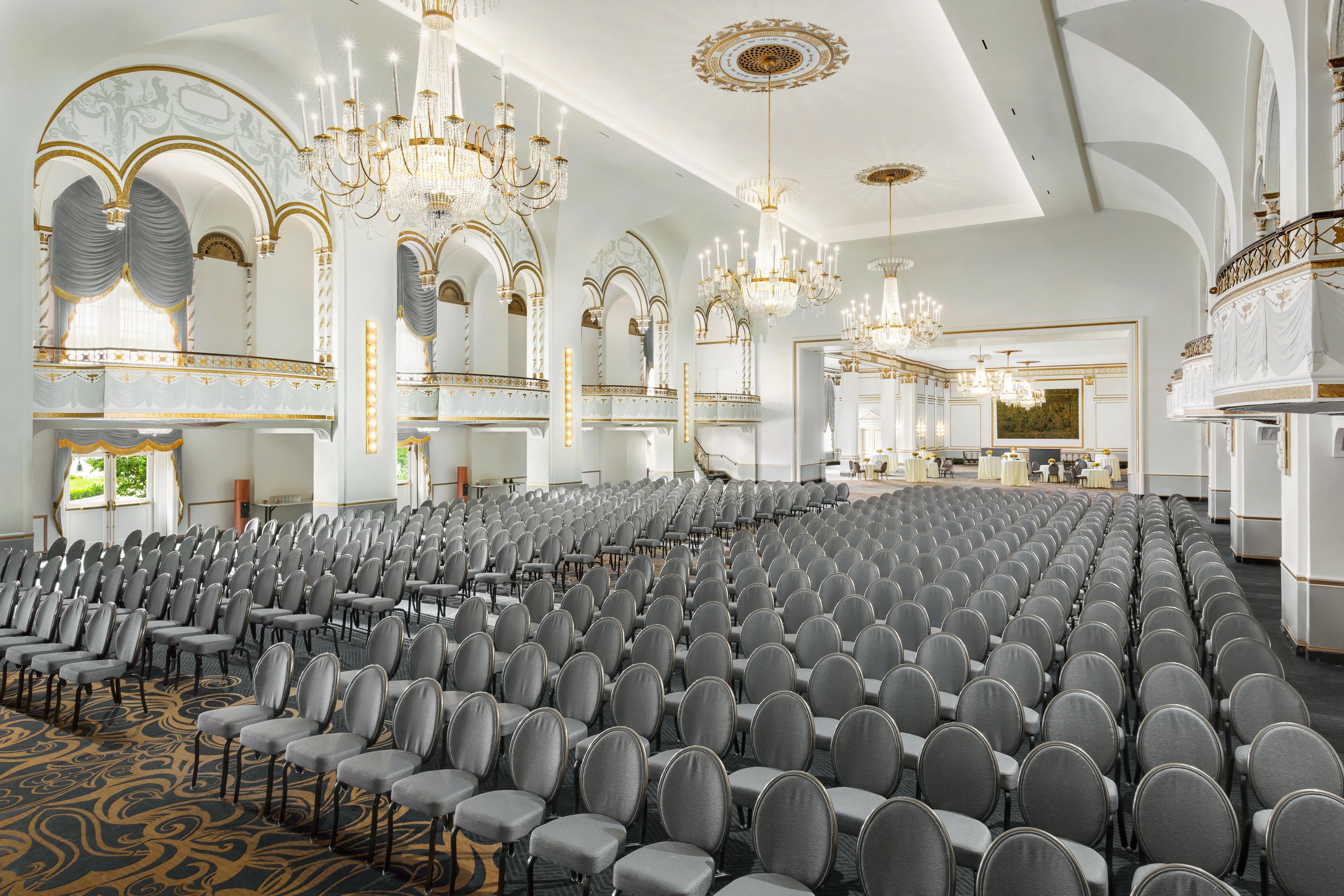 A large hall with rows of chairs, ornate chandeliers, and a grand staircase.