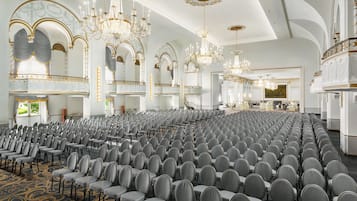 A large hall with rows of chairs, ornate chandeliers, and a grand staircase.