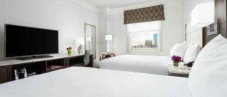 Habitación de hotel con dos camas, un televisor de pantalla plana, un escritorio con lámpara y ventana con vista a la ciudad.