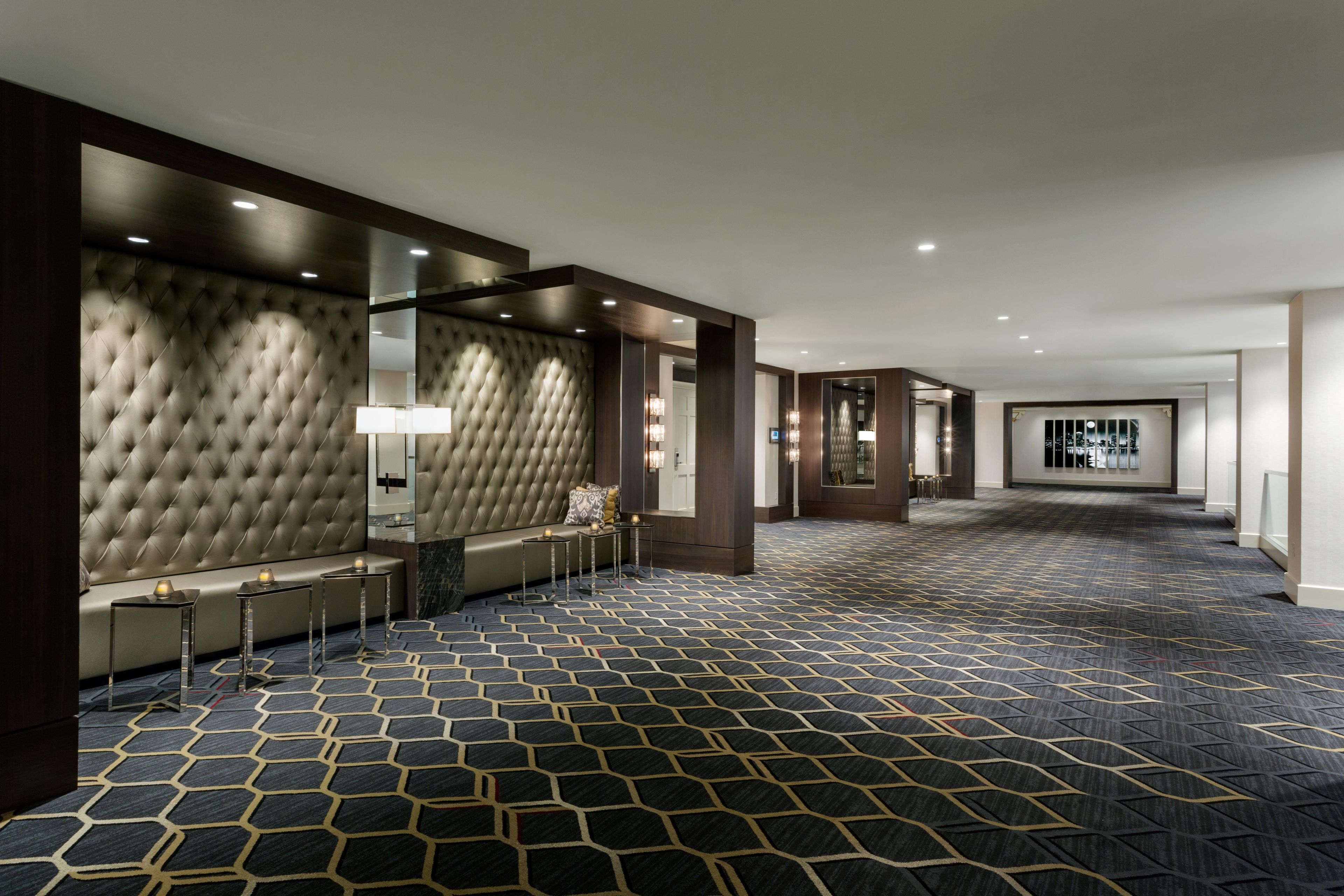 Un couloir d’hôtel avec un tapis à motifs, un coin salon agrémenté d’un mur capitonné et un miroir qui reflète la pièce.