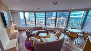 A modern living room with a curved sofa, round coffee table, and a dining area with chairs. Large windows offer a view of the cityscape.