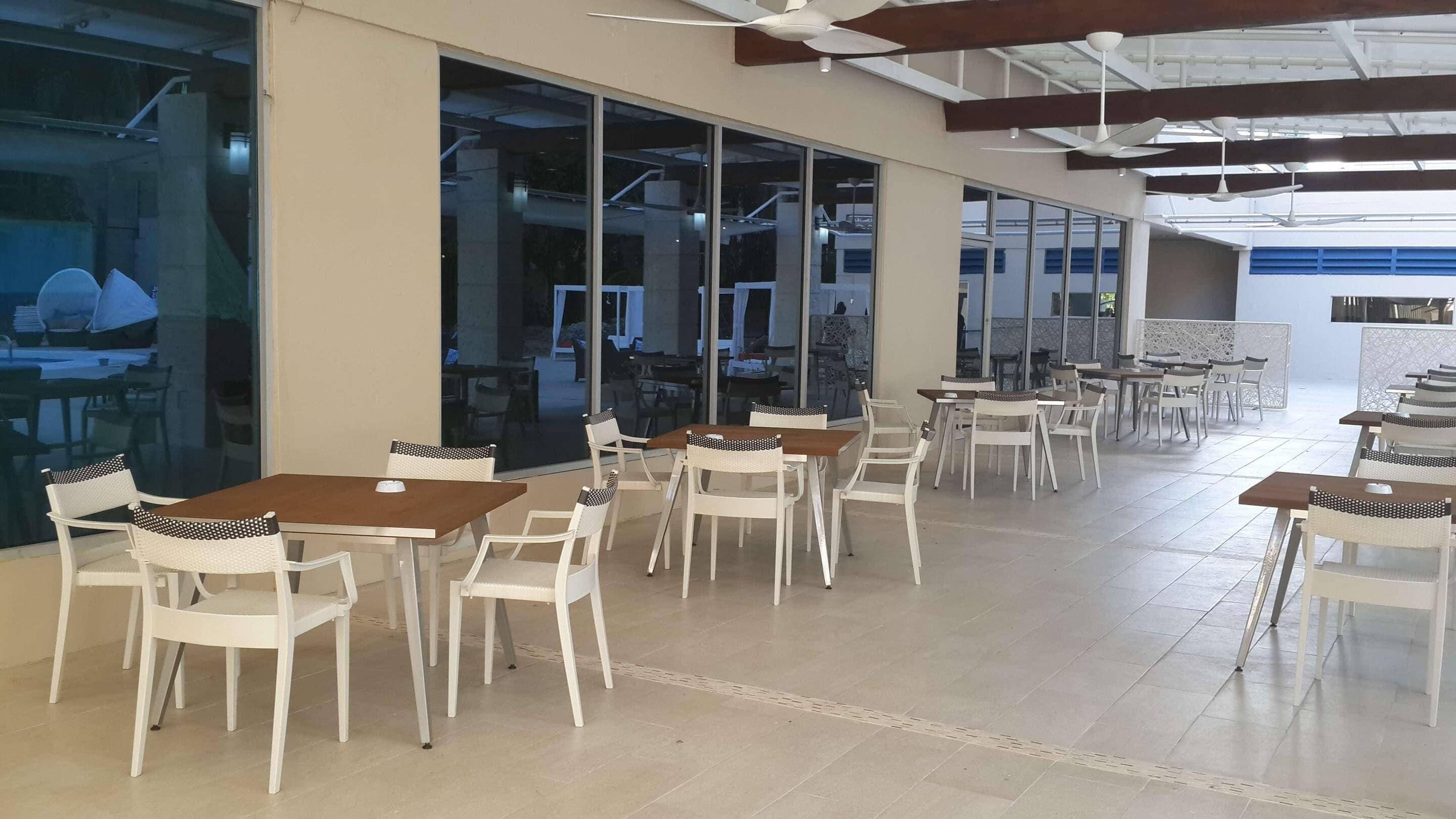 Een ruime eethoek binnen met meerdere tafels en stoelen, grote ramen en plafondventilatoren.
